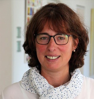 Lächelnde Frau mit braunen Haaren und Brille, die einen weiß-gepunkteten Schal mit einem weißen Shirt trägt und in einem Flur steht