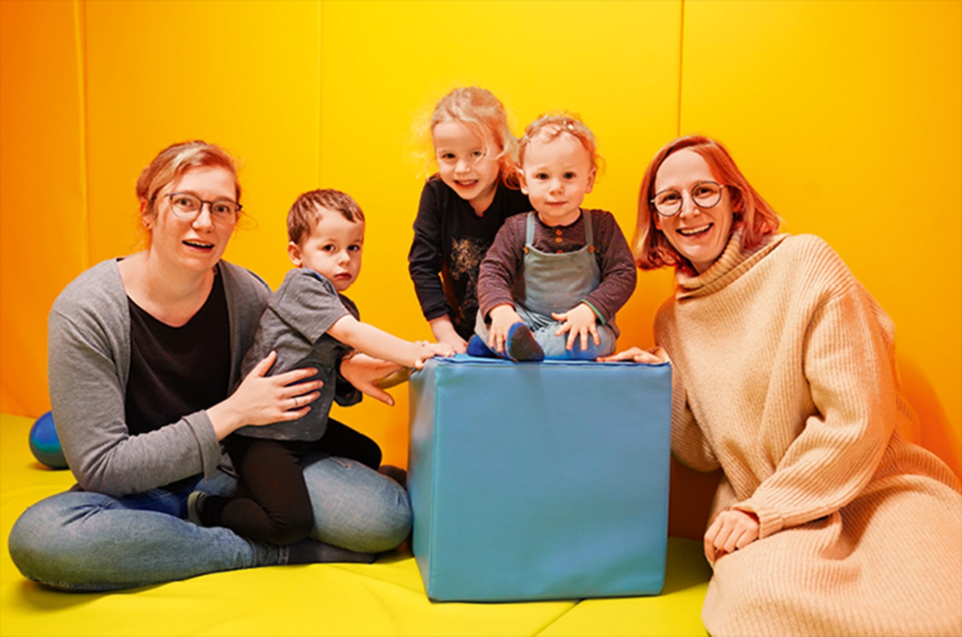 Zwei Frauen sitzen mit drei Kindern in einem gelb-orange ausgekleideten Time-out-Raum einer Kurklinik in Goch. Mitten im Raum steht ein blauer Sitzwürfel.