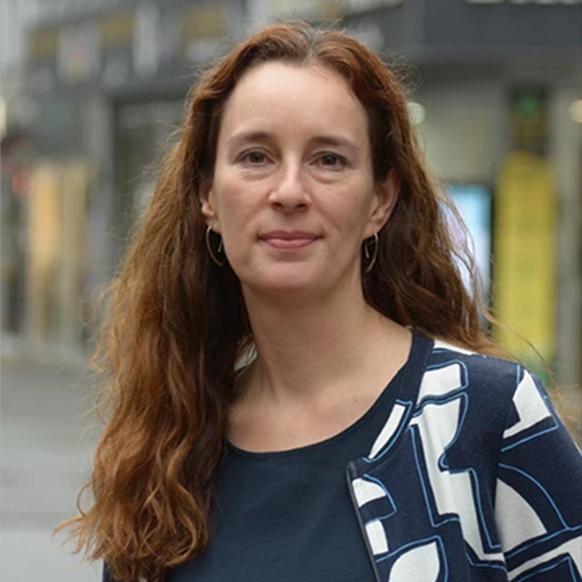 Sanft lächelnde Frau mit langen braunen Haaren, die ein blaues Shirt und eine eine blau-weiße Jacke trägt. Sie steht in der Fußgängerzone der Essener Innenstadt.