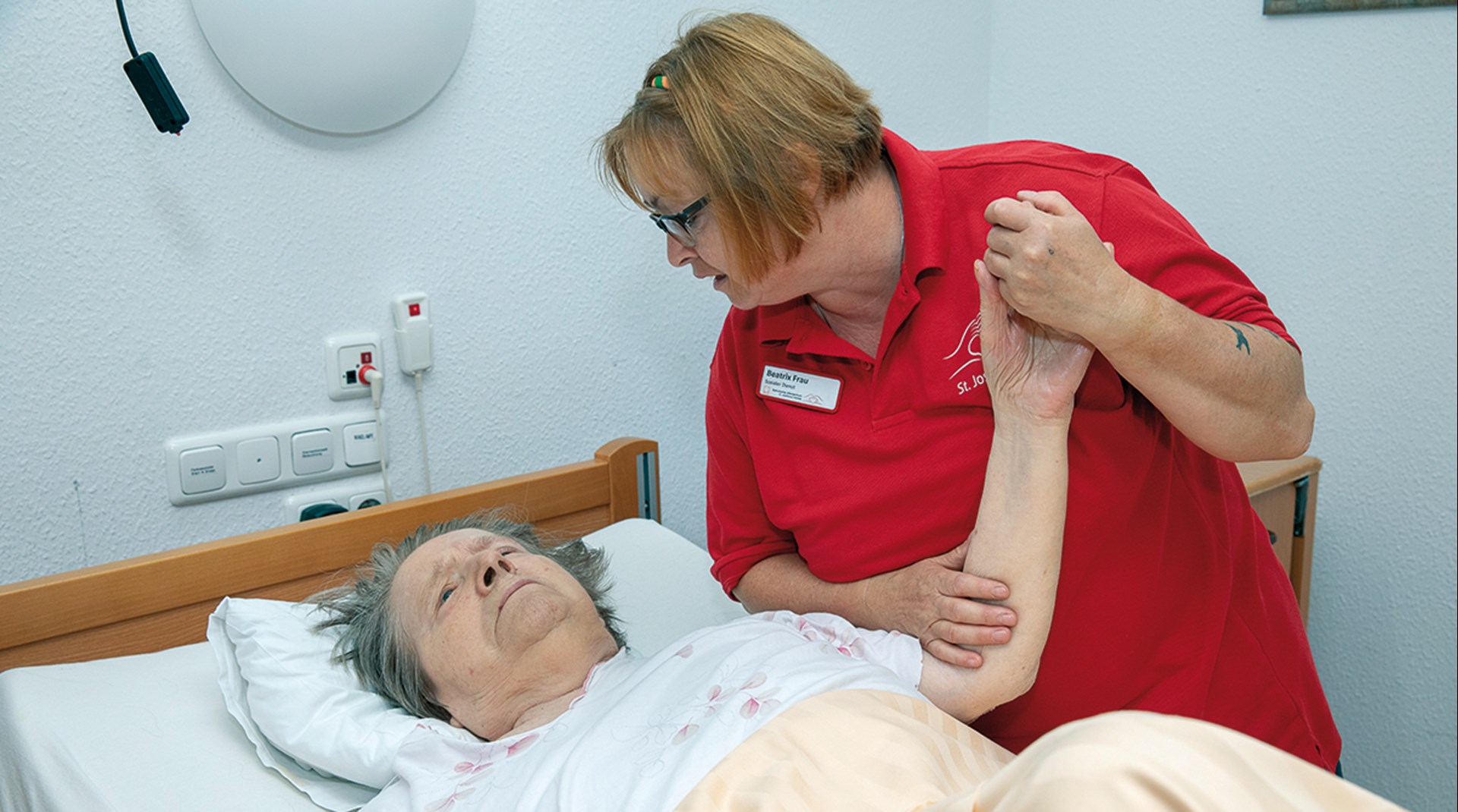 Eine Pflegerin, die an einem Pflegebett steht und den ausgestreckten Arm einer im Bett liegenden Seniorin hält