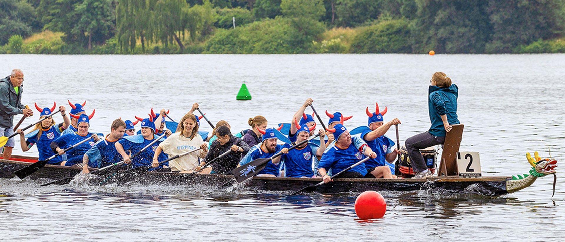 Ein voll besetztes Drachenboot der "Wikinger vom Hellweg-Express", dass auf dem Essener Baldeneysee fährt. Die meisten Teammitglieder tragen ein blaues Trikot mit einem blau-orangen Wikingerhelm.