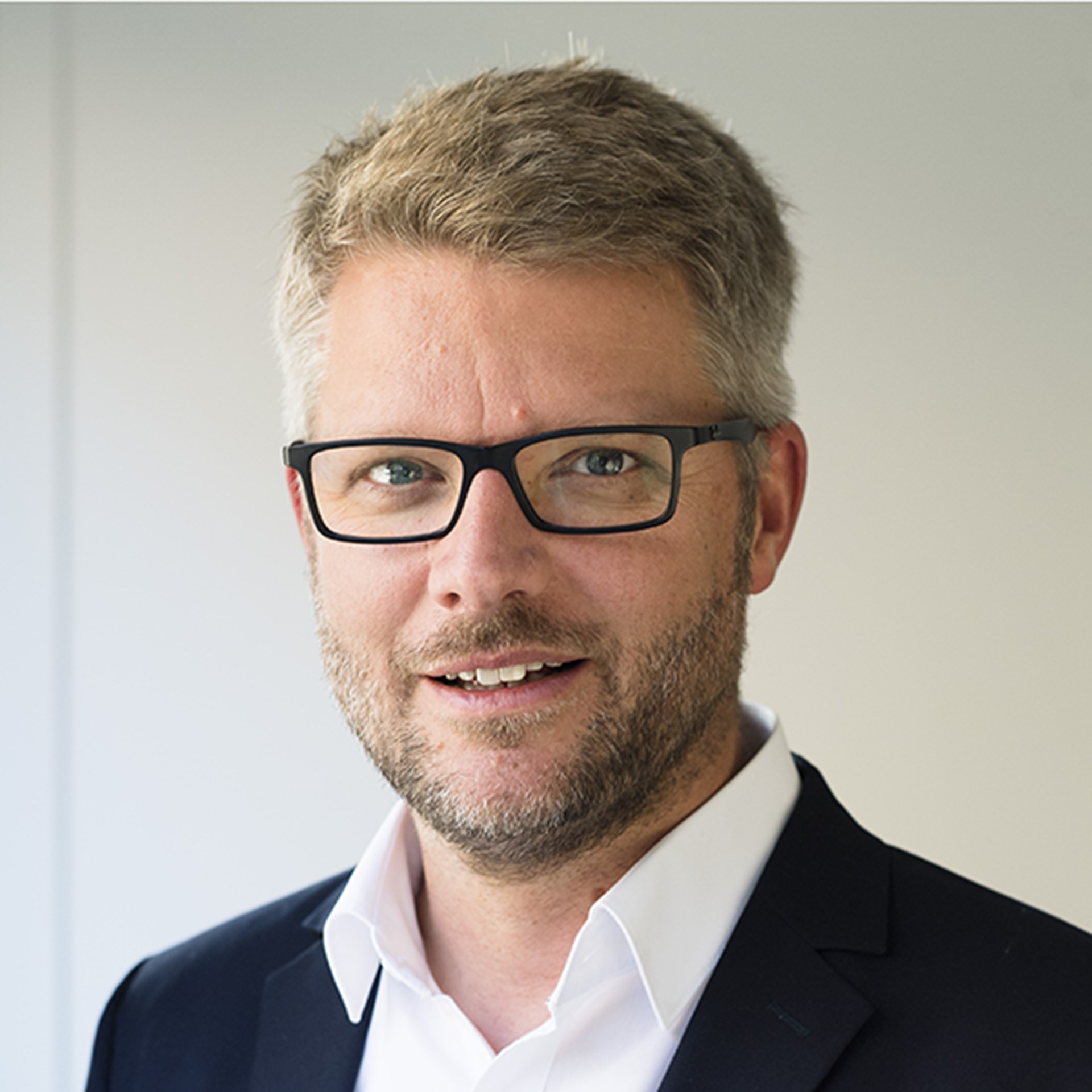 Lächelnder Mann mit blonden Haaren, Brille und Bart, der ein weißes Hemd und eine schwarze Anzugjacke trägt