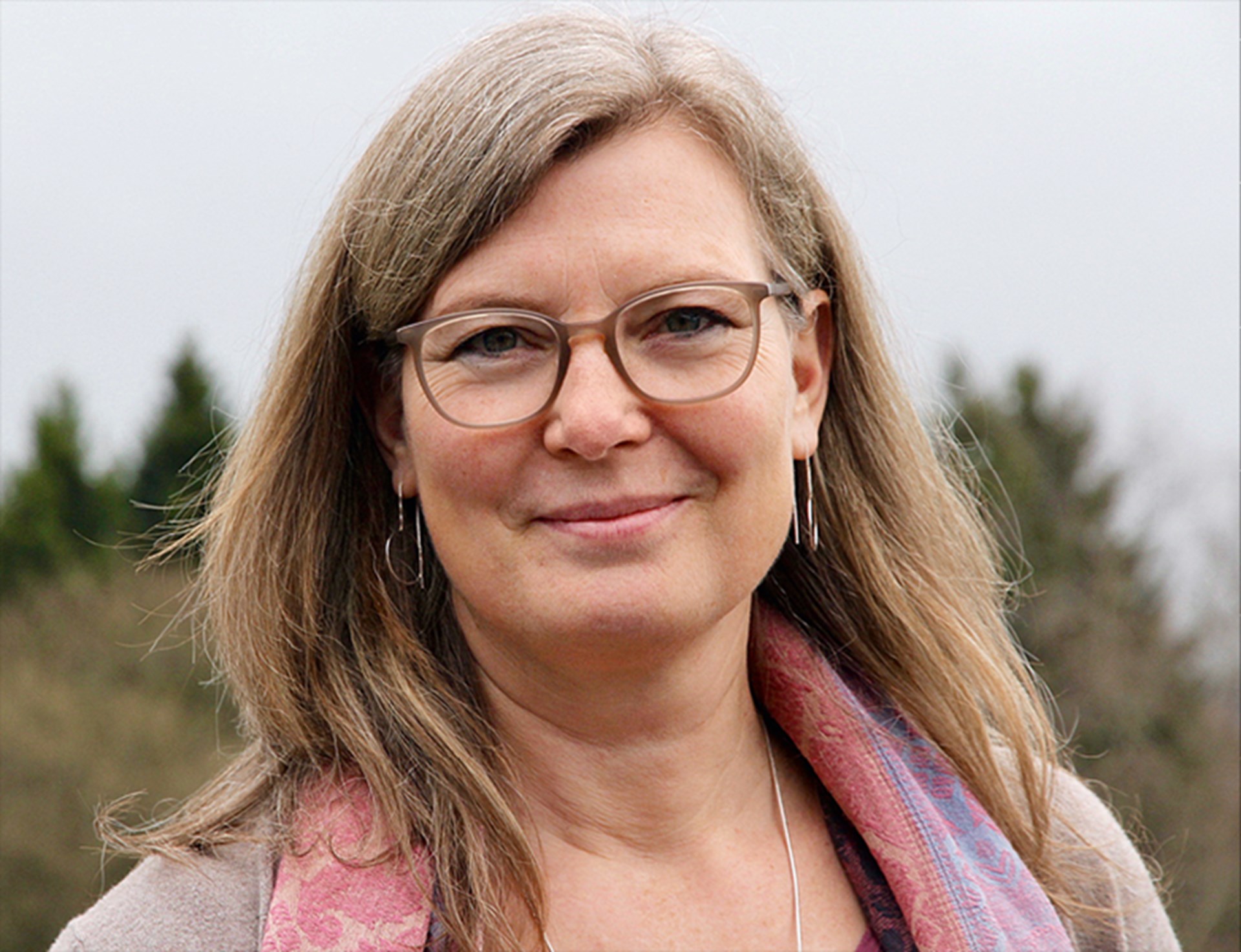Sanft lächelnde Frau mit langen, blonden Haaren und einer braunen Brille, die einen rötlichen Schal mit einer hellen Jacke trägt und vor einem Feld steht