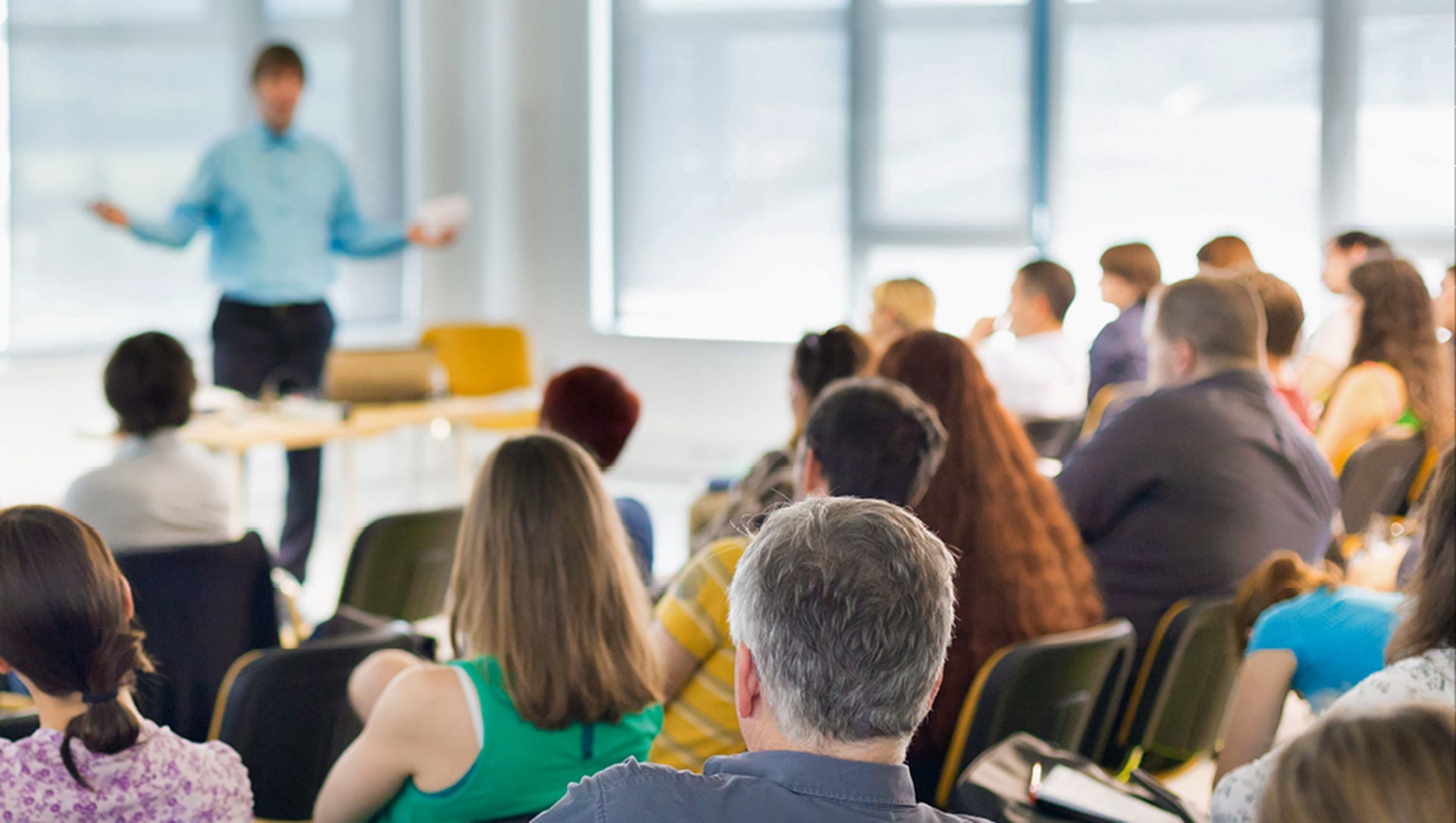 Ein Publikum, dass in einem Schulungsraum in Stuhlreihen vor einem Tisch befindet, an dem ein Mann einen Vortrag hält