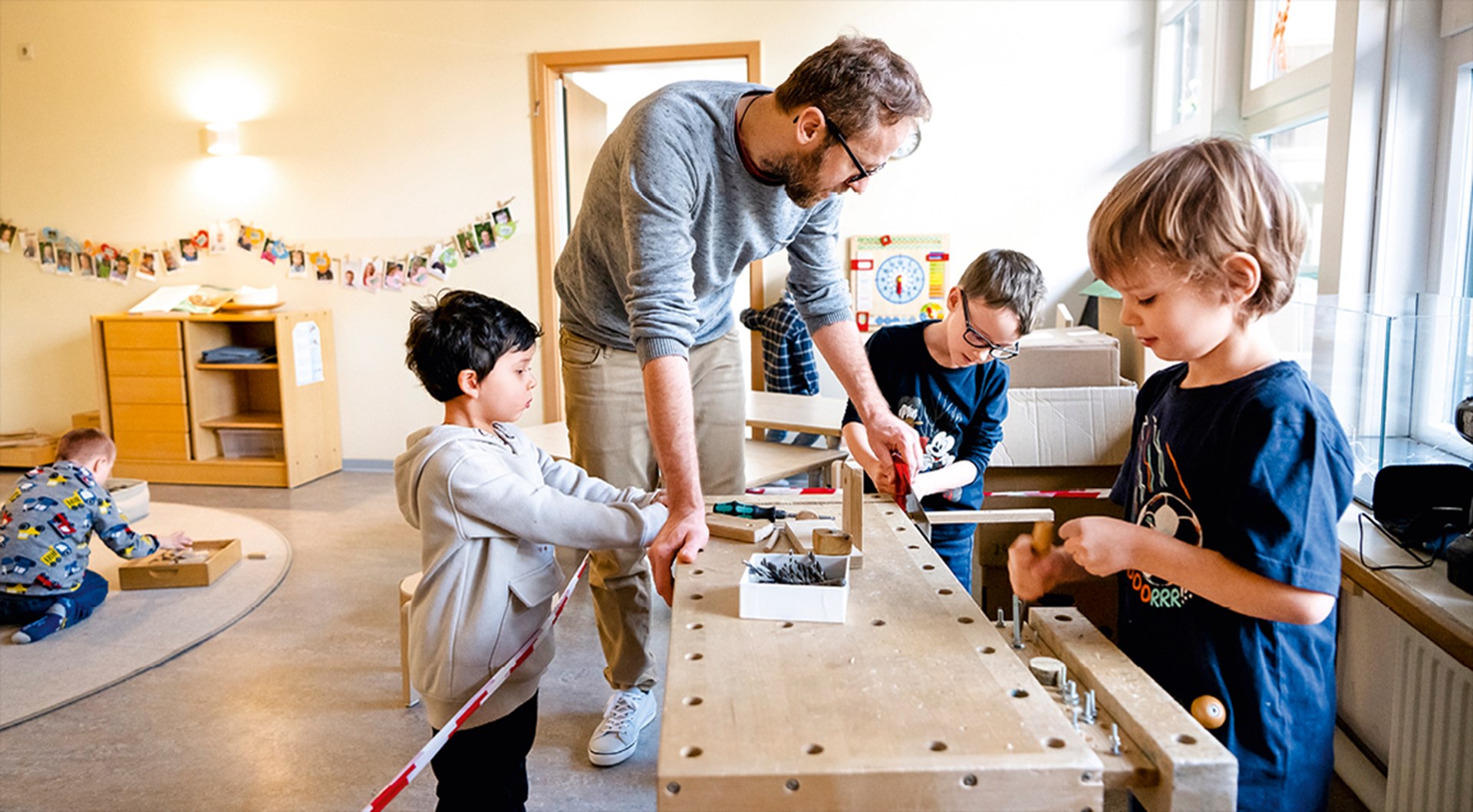 Ein Erzieher, der in einer Kita mit drei Kindern an eine Katapult arbeitet. Im Hintergrund spielt ein weiteres Kind auf einem Teppich.