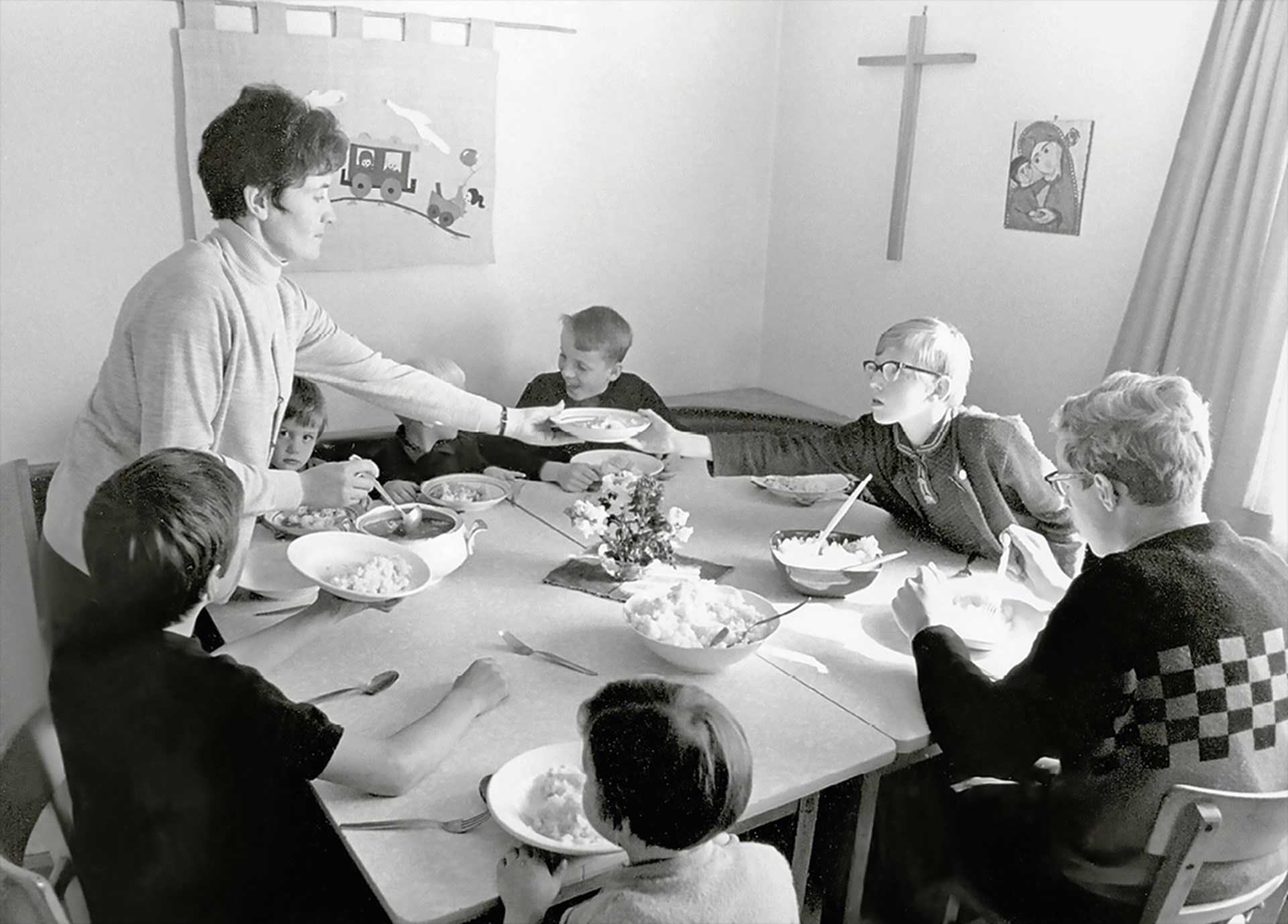 Sieben Jungen sitzen zusammen um einen aus zwei Tischen zusammengestellten Esstisch und nehmen eine Mahlzeit ein. Eine Betreuerin, die aus einem Kochtopf etwas zu Essen schöpft, reicht einem Jungen einen gefüllten Teller herüber. Das Foto ist in schwarz-weiß.