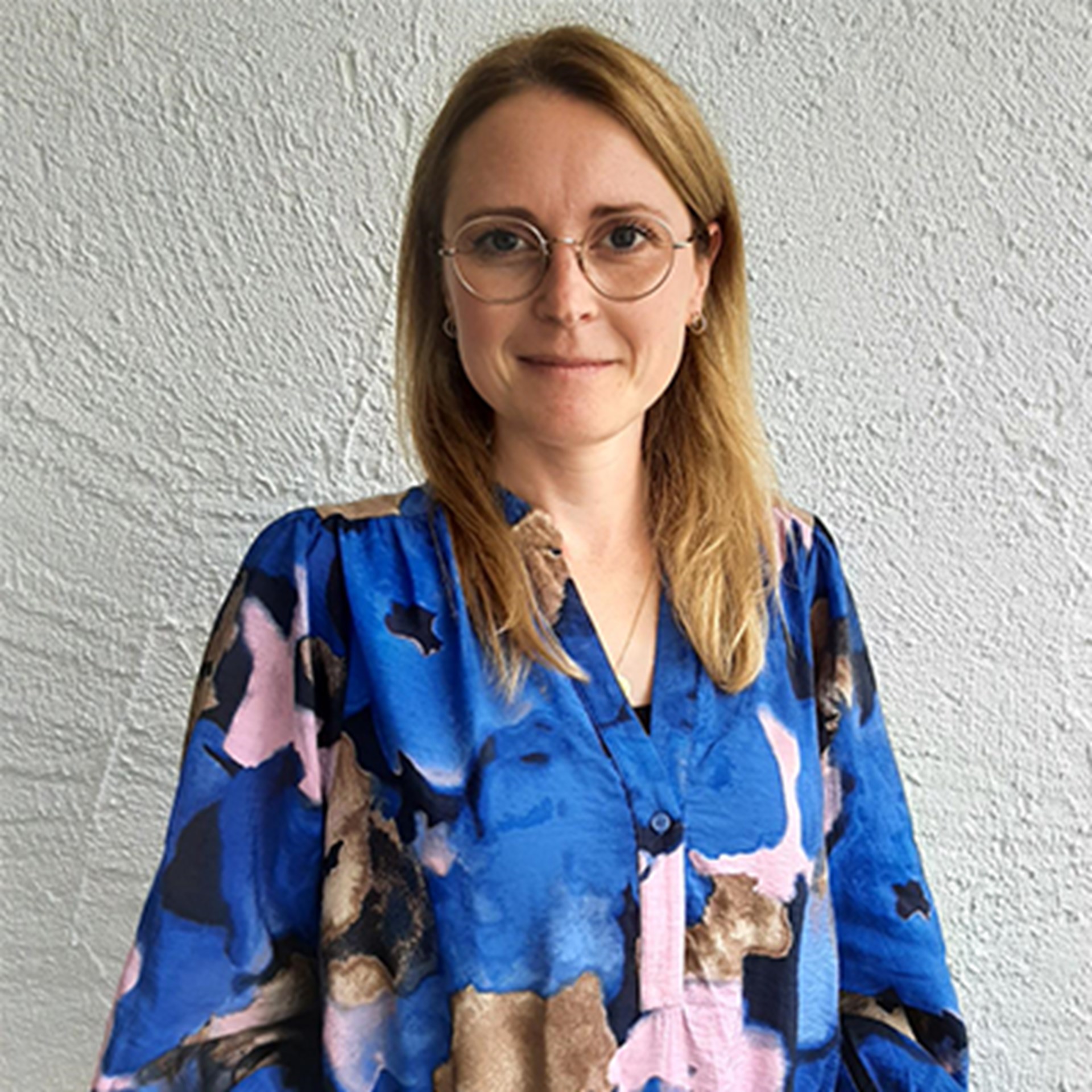 Frau mit langen, blonden Haaren und einer Brille, die eine blaue Bluse mit buntem Muster trägt