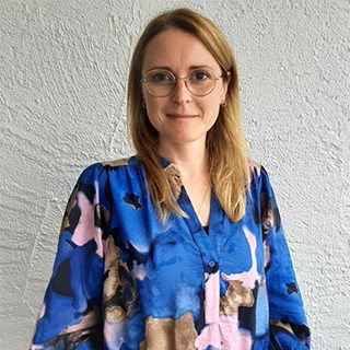 Frau mit langen, blonden Haaren und einer Brille, die eine blaue Bluse mit buntem Muster trägt