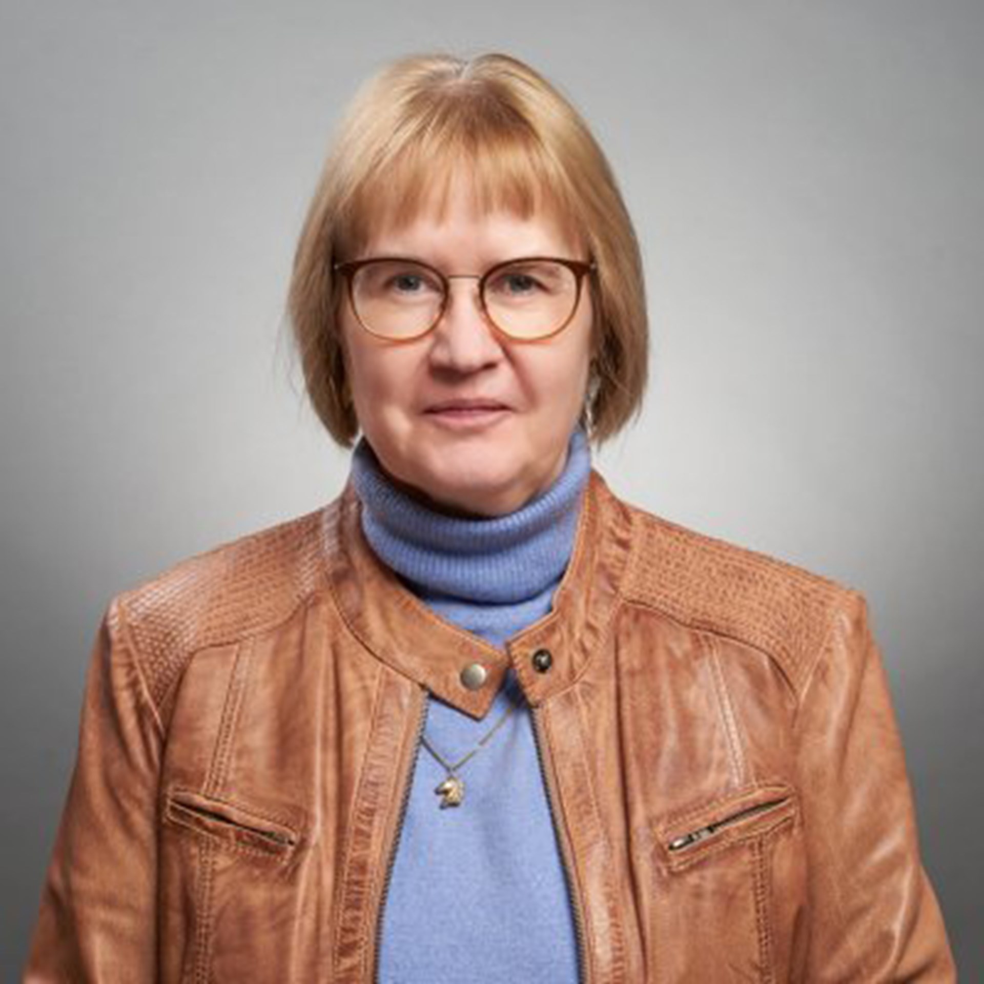 Frau mit rötlichen Haaren und Brille, die einen hellblauen Rollkragenpullover und eine hell-braune Lederjacke trägt