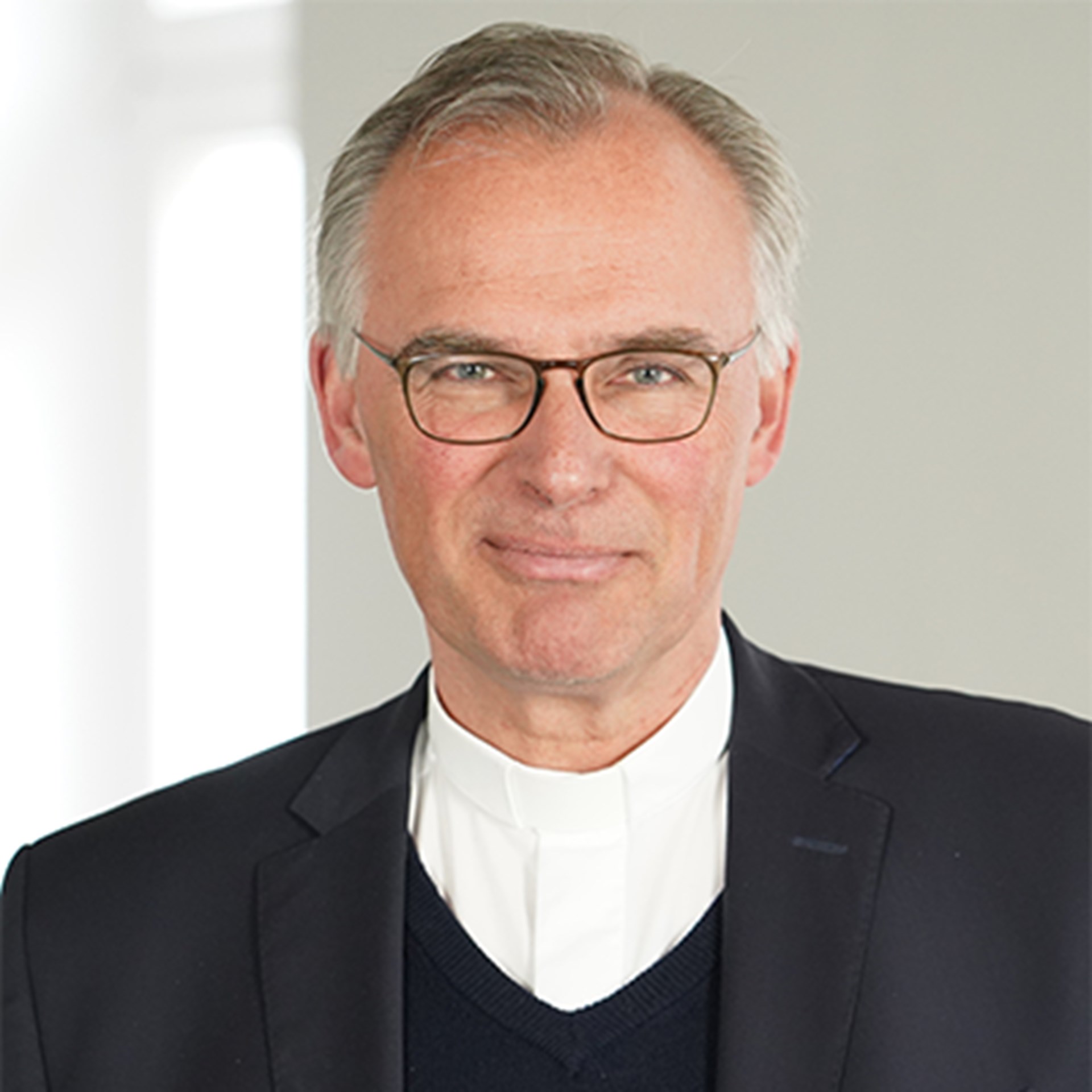 Lächelnder Mann mit grauen Haaren und Brille, der ein weißes Hemd, einen schwarzen Pullover und eine schwarze Anzugjacke trägt