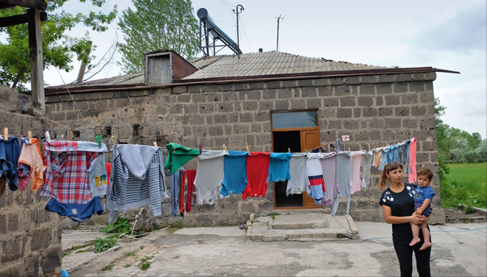 Ein Steinhaus in Armenien mit einer voll behangenen Wäscheleine, vor der eine Frau steht, die ein Kind auf dem Arm hat
