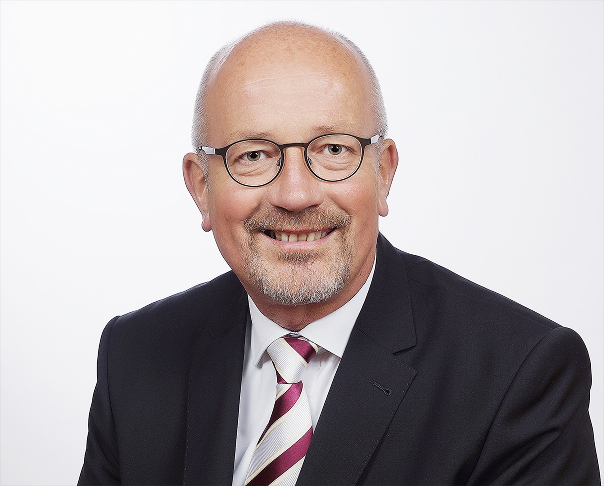 Lächelnder Mann mit grauen Haaren, Halbglatze, Brille und Bart, der ein weißes Hemd, eine rot-weiß-gestreifte Krawatte und eine schwarze Anzugjacke trägt