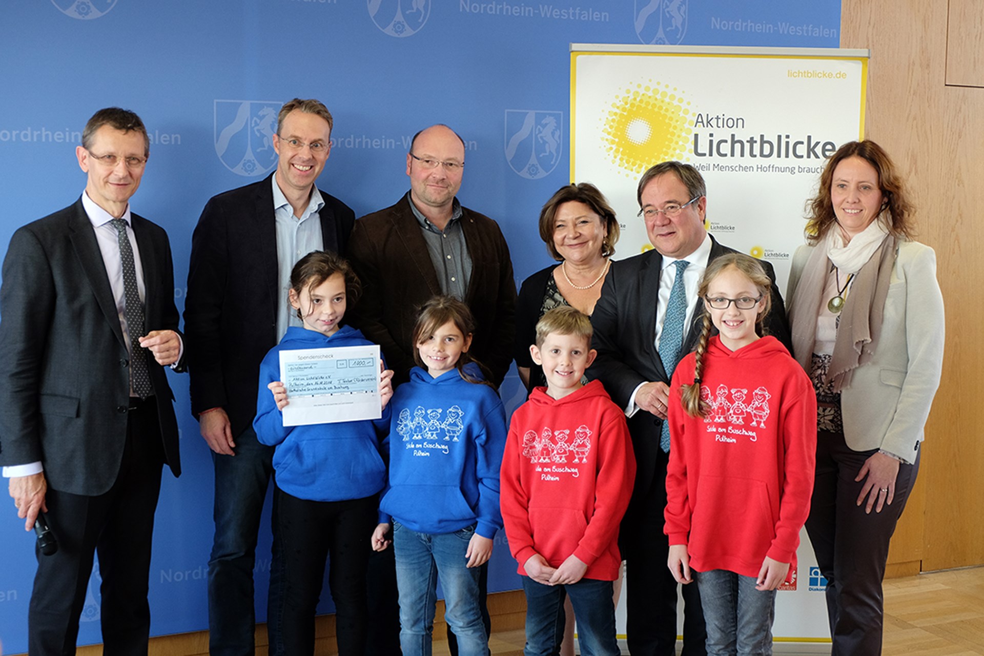 Ein Gruppenfoto mit vier Schüler/innen der kath. Grundschule am Buschweg in Pulheim und Vertreter/innen der Aktion Lichtblicke. Eine Schülerin hält einen Scheck in den Händen.