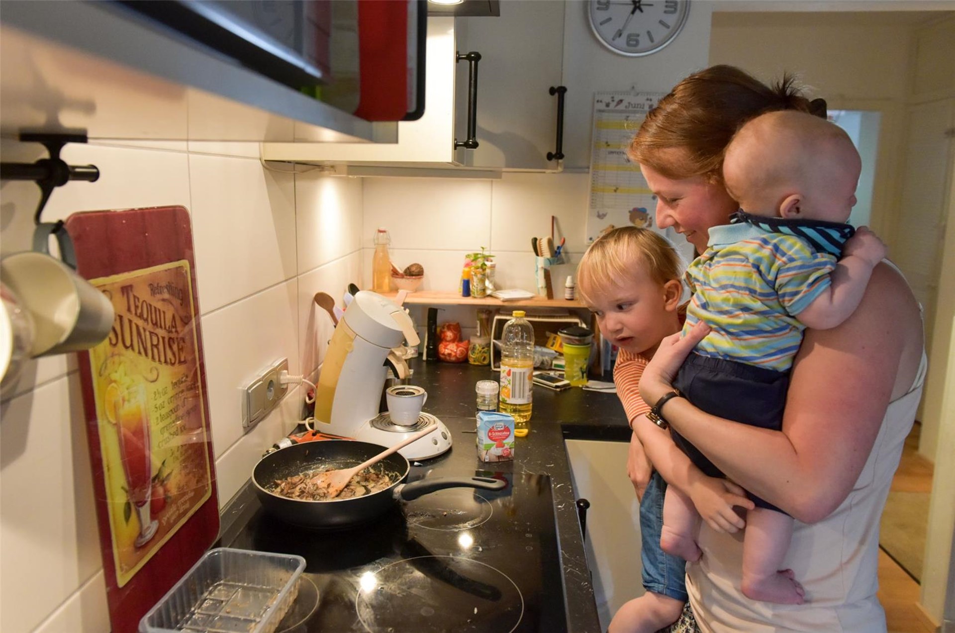 Mutter hält ein Kleinkind und ein Baby auf dem Arm während sie kocht