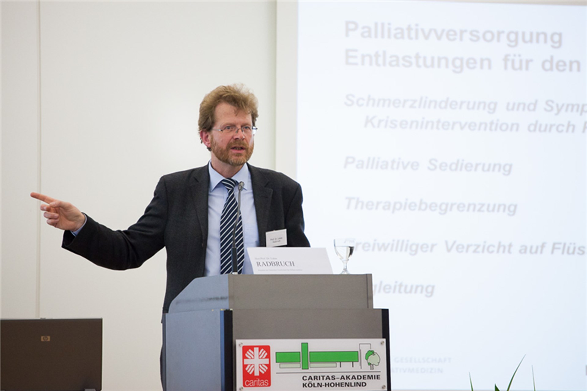 Ein Mann steht beim Akademietag vor einem Pult und hält eine Rede. Er deutet dabei mit seinem rechten Zeigefinger zur Seite. Hinter ihm ist eine Leinwand zu sehen.