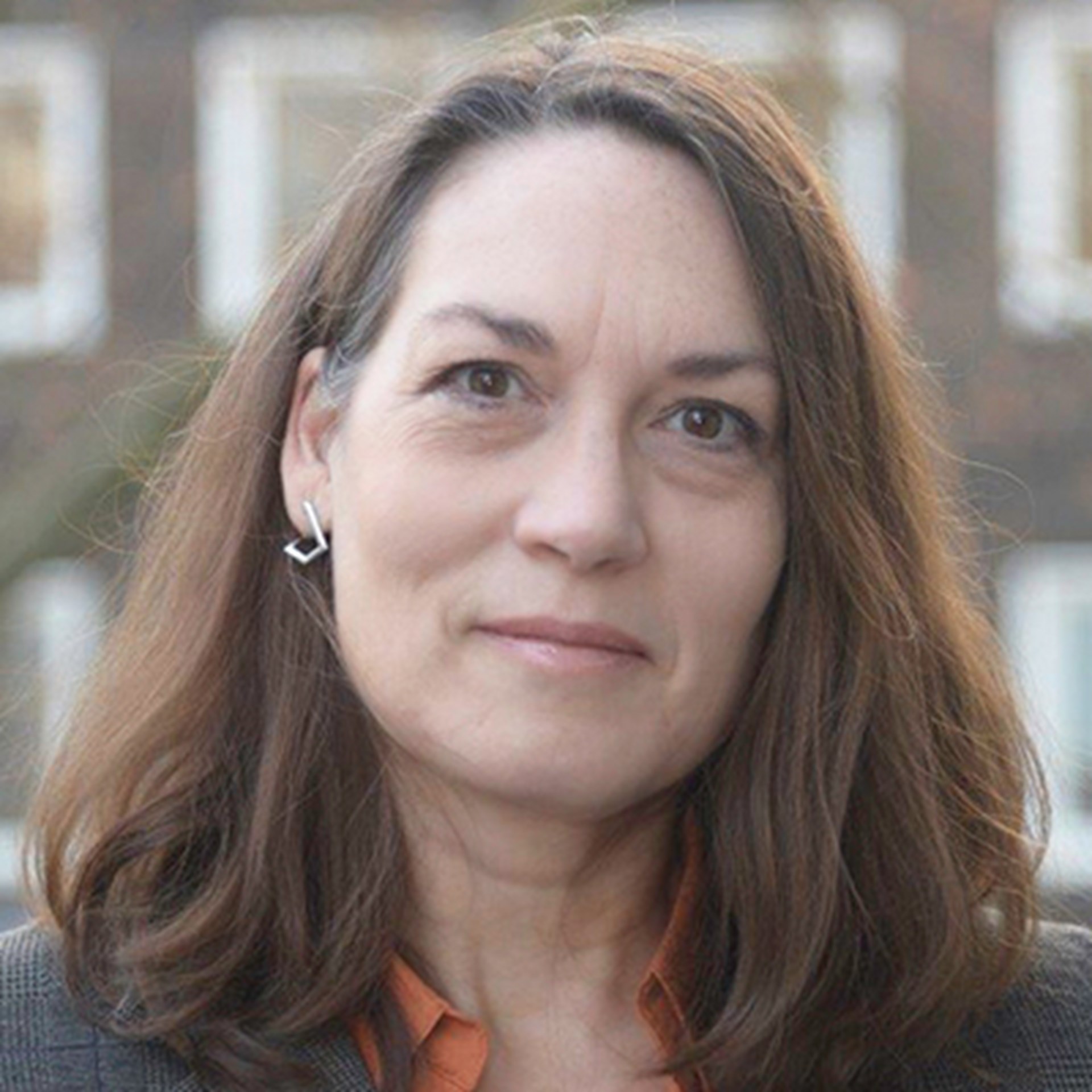 Sanft lächelnde Frau mit langen, braunen Haaren, die ein orangenes Hemd mit einer grauen Anzugjacke trägt