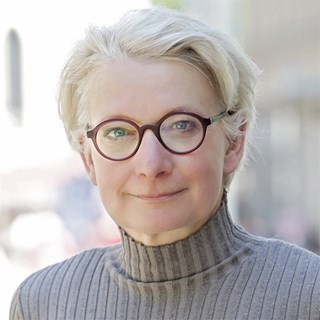 Lächelnde Frau mit hellblonden Haaren und Brille, die einen grauen Rollkragenpullover trägt und sich in der Fußgängerzone der Essener Innenstadt befindet