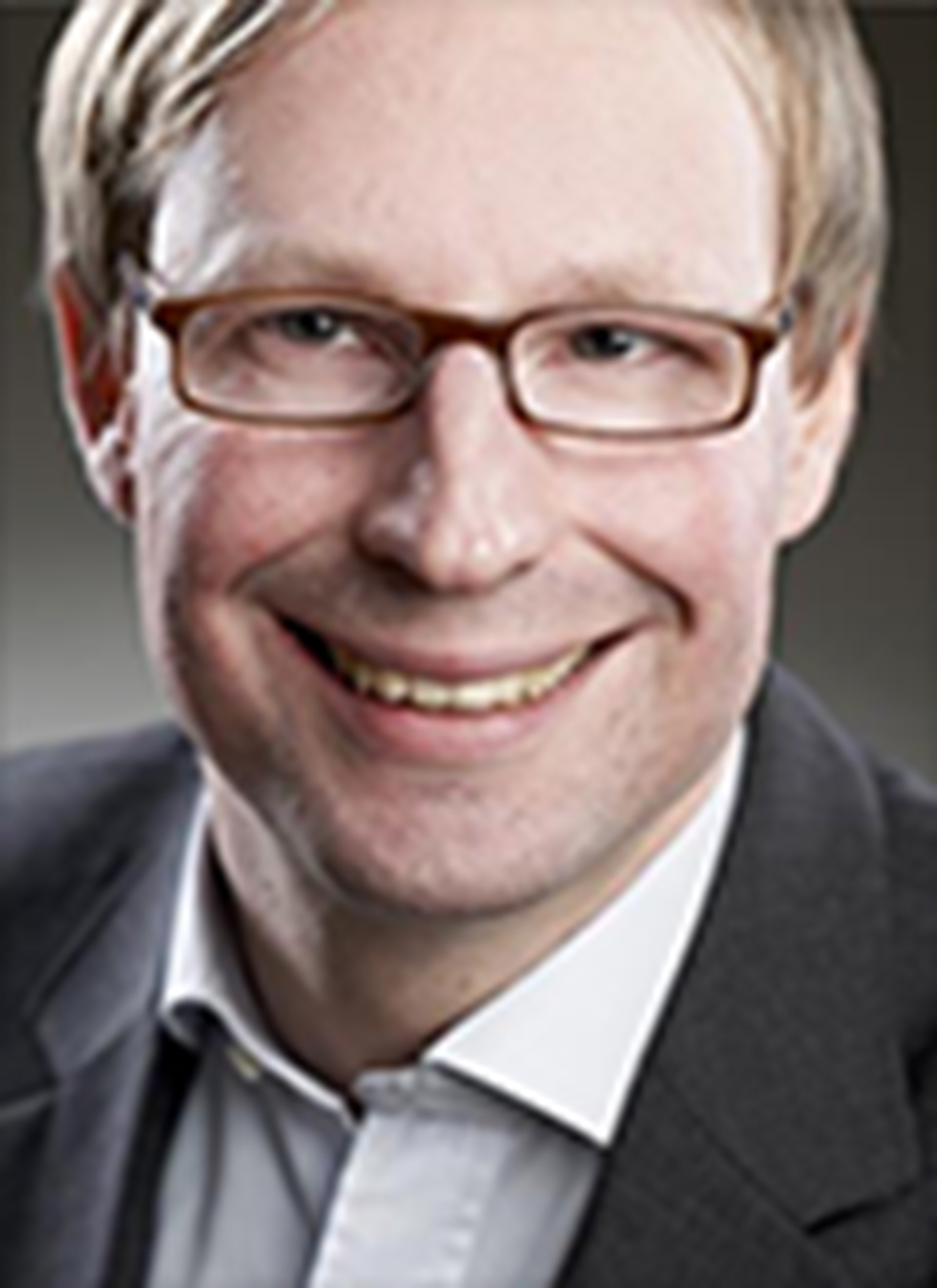 Lächelnder Mann mit blonden Haaren und Brille, der ein hellgraues Hemd und eine graue Anzugjacke trägt