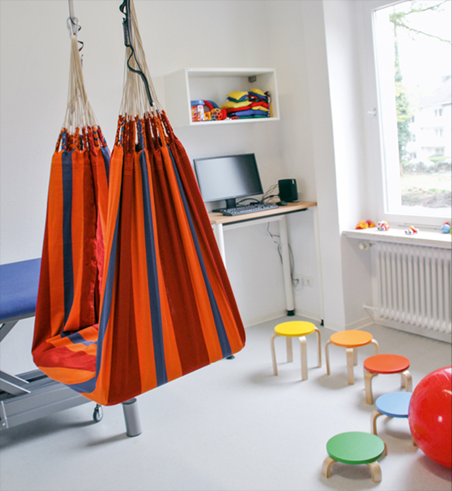 Ein Raum im Diagnose- und Therapiezentrum des Franz Sales Hauses, der unter anderem mit einer Hängematte, einer Liege, einigen Hockern und einem Bildschirmarbeitsplatz ausgestattet ist.