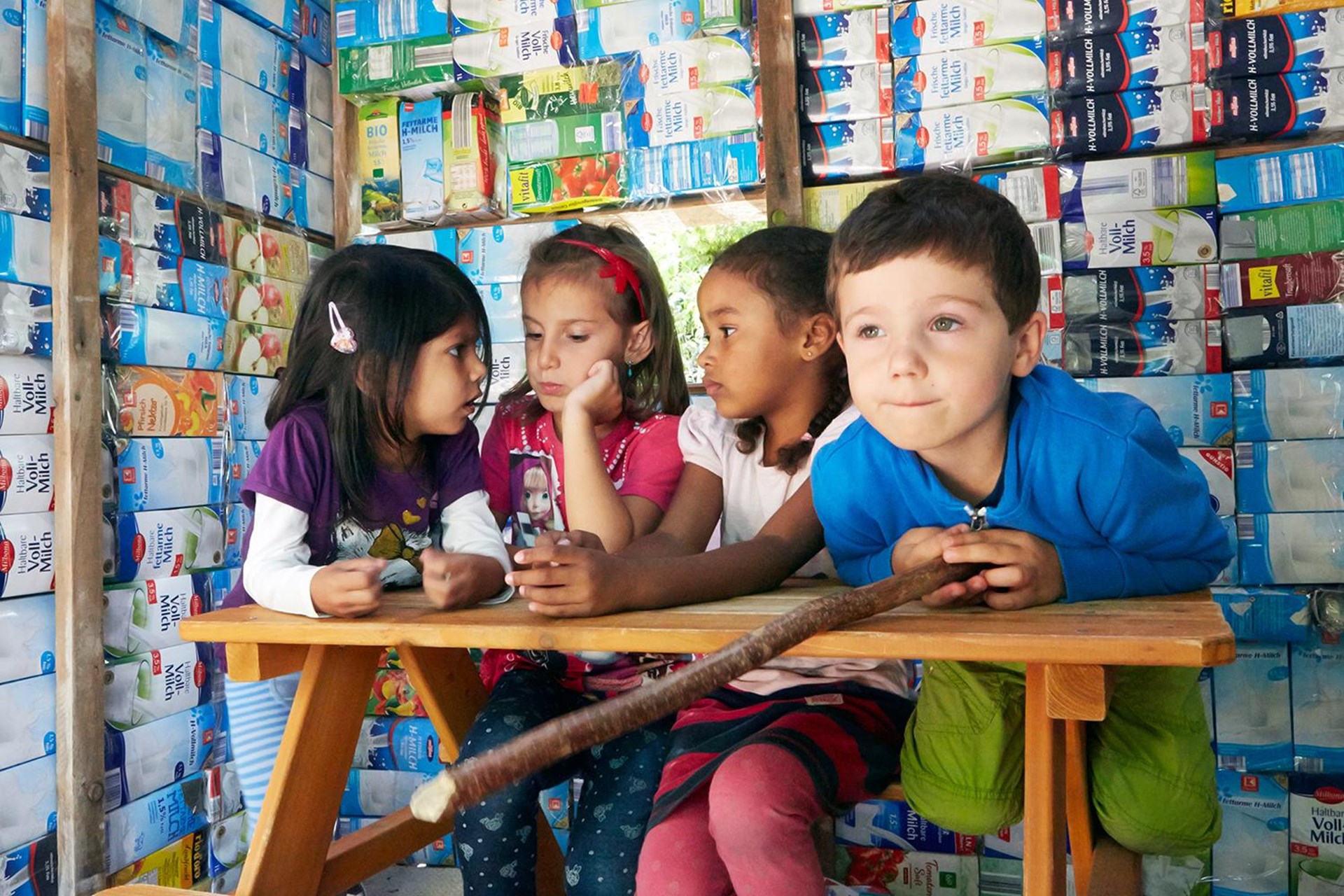 Vier Kinder sitzen an einem Tisch, im Hintergrund eine künstlerische Wand aus Milchtüten.