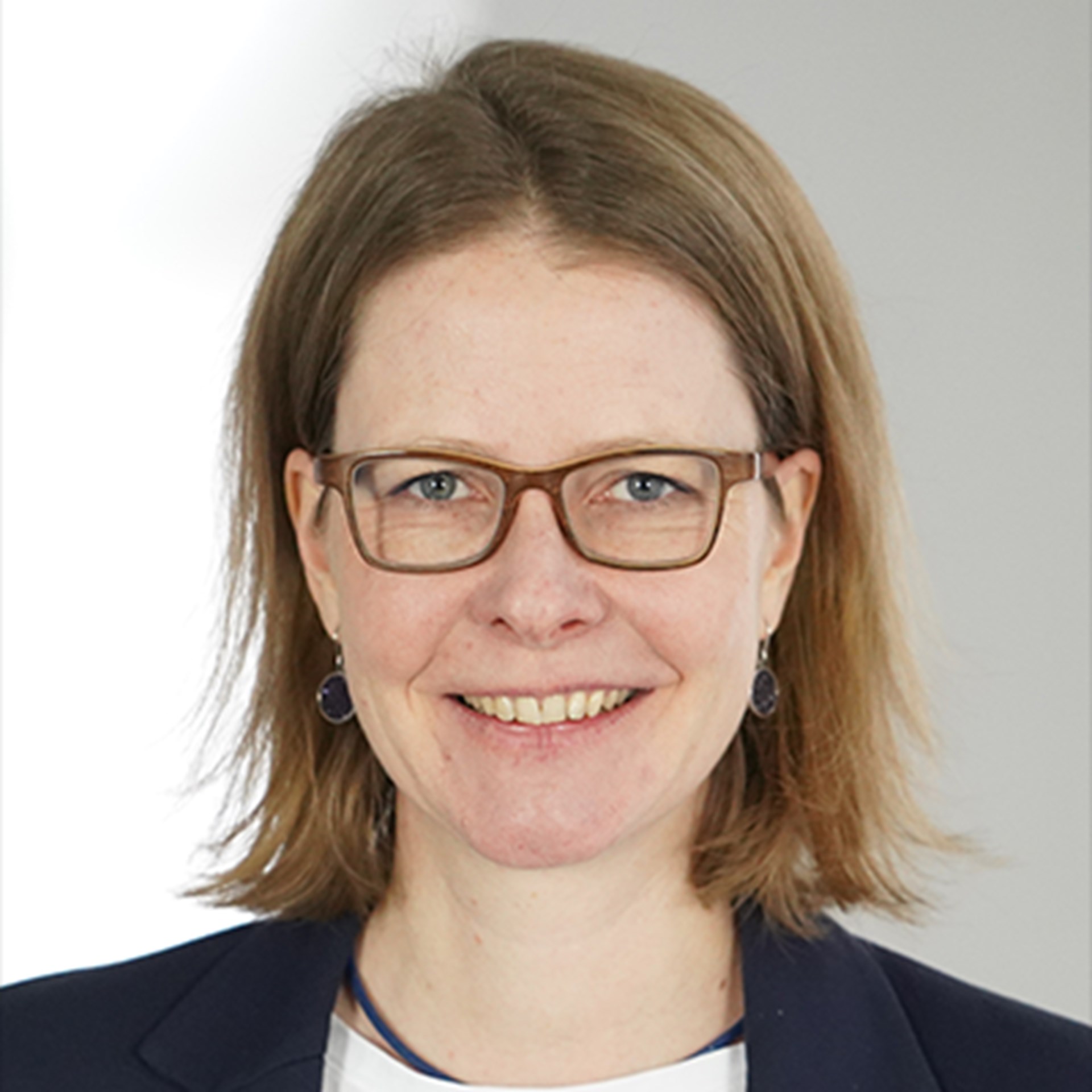 Lächelnde Frau mit langen, braunen Haaren und blauer Brille, die ein weißes Shirt und eine blaue Anzugjacke trägt