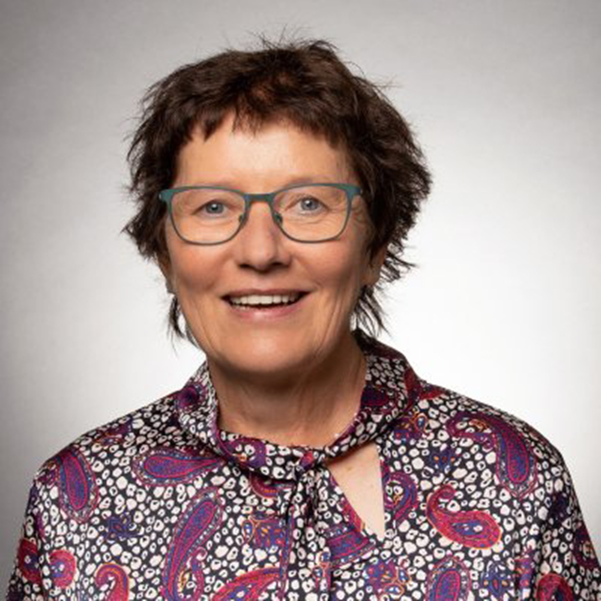 Lächelnde Frau mit braunen Haaren, einer Brille und einem bunten Oberteil