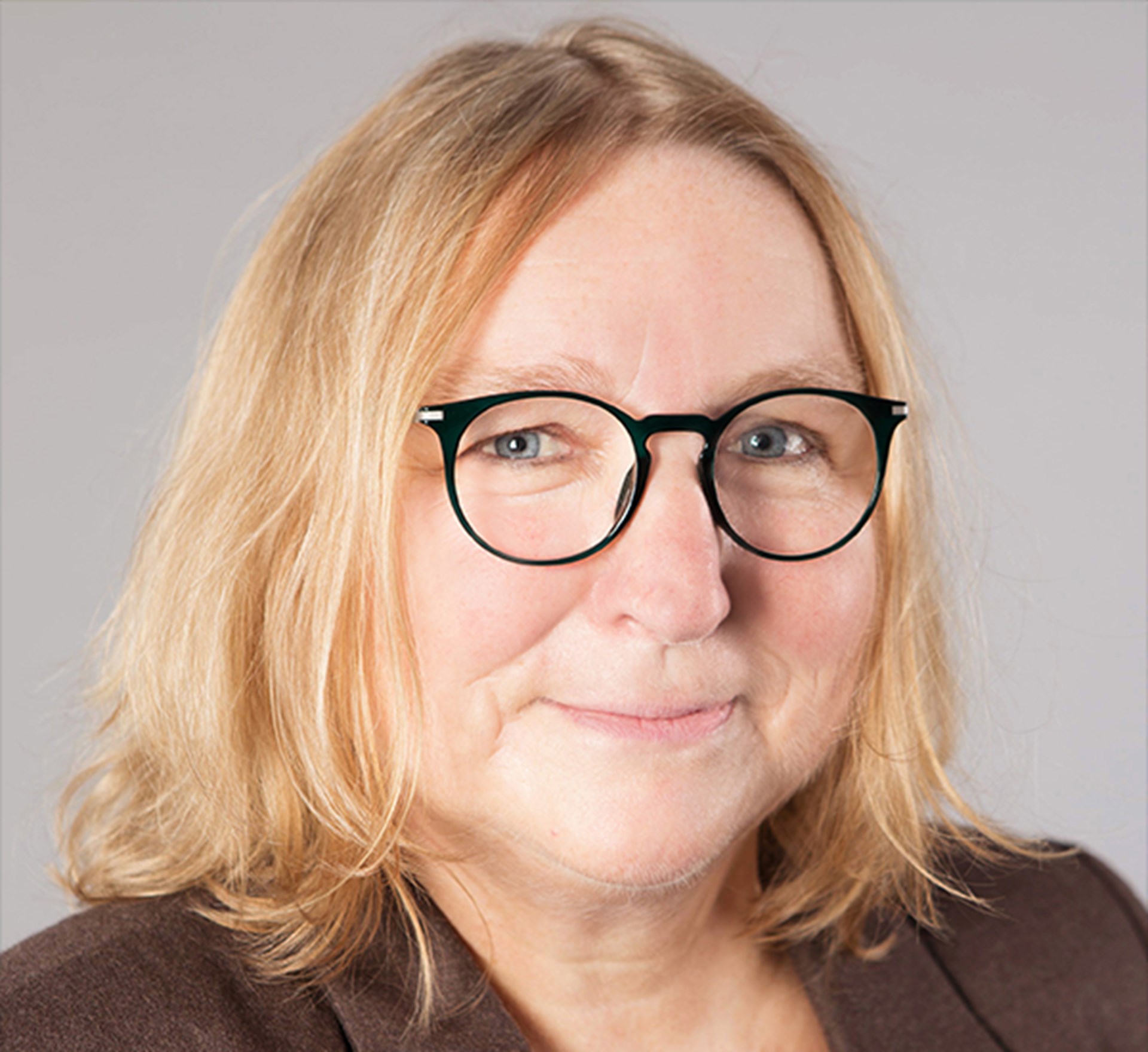 Lächelnde Frau mit hellblonden Haaren und Brille, die braune Kleidung trägt