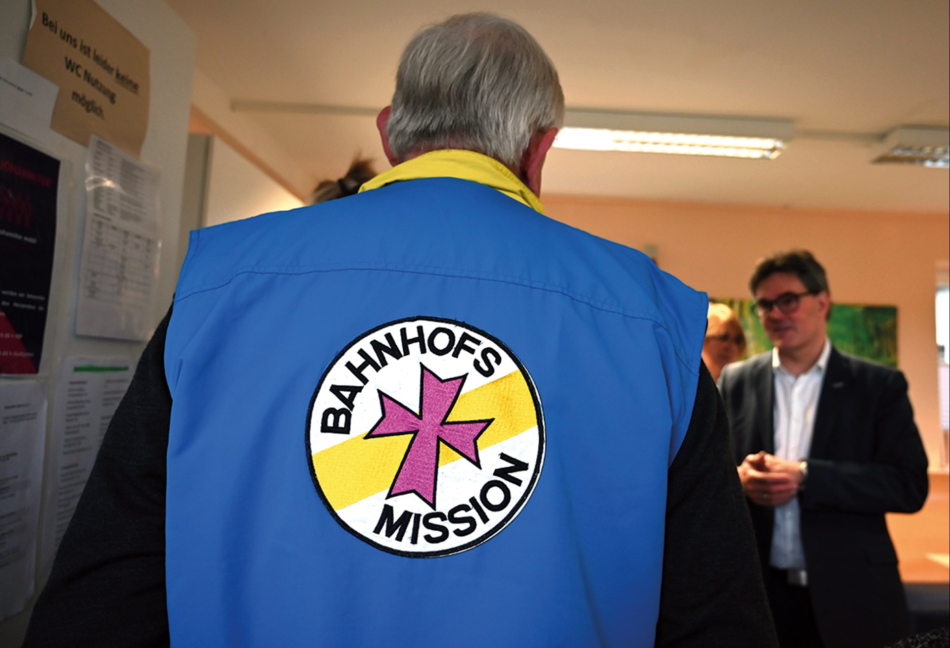 NRW-Sozialminister Karl-Josef Laumann steht in einem Raum vor einem anderen Mann und trägt dabei eine blaue-gelbe Weste mit dem Logo der Bahnhofsmission. Er steht mit dem Rücken zur Kamera.