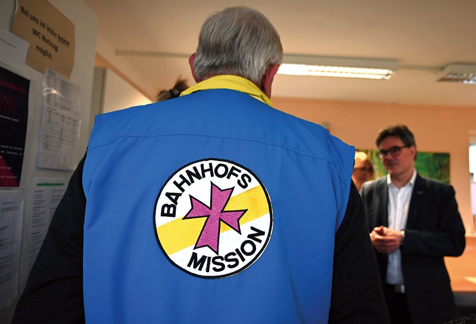 NRW-Sozialminister Karl-Josef Laumann steht in einem Raum vor einem anderen Mann und trägt dabei eine blaue-gelbe Weste mit dem Logo der Bahnhofsmission. Er steht mit dem Rücken zur Kamera.