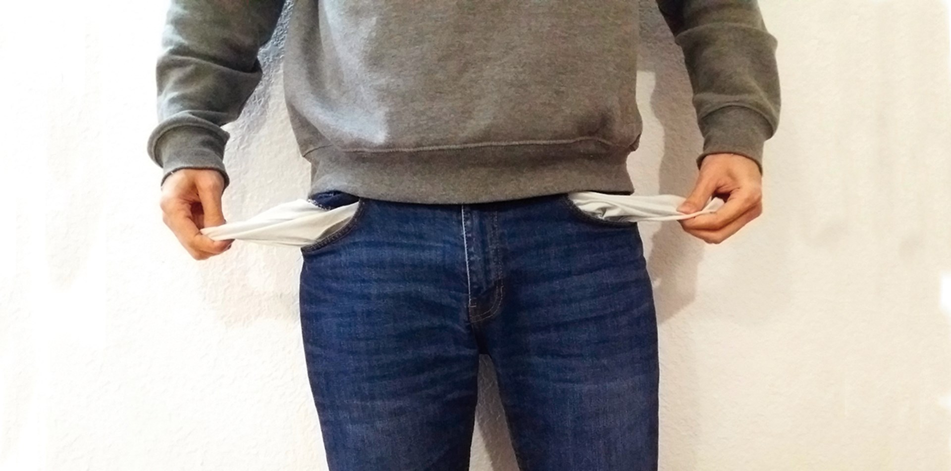 Ein Mann im grauen Pullover und blauer Jeans, der die Innenseite seiner leeren Hosentaschen herauszieht
