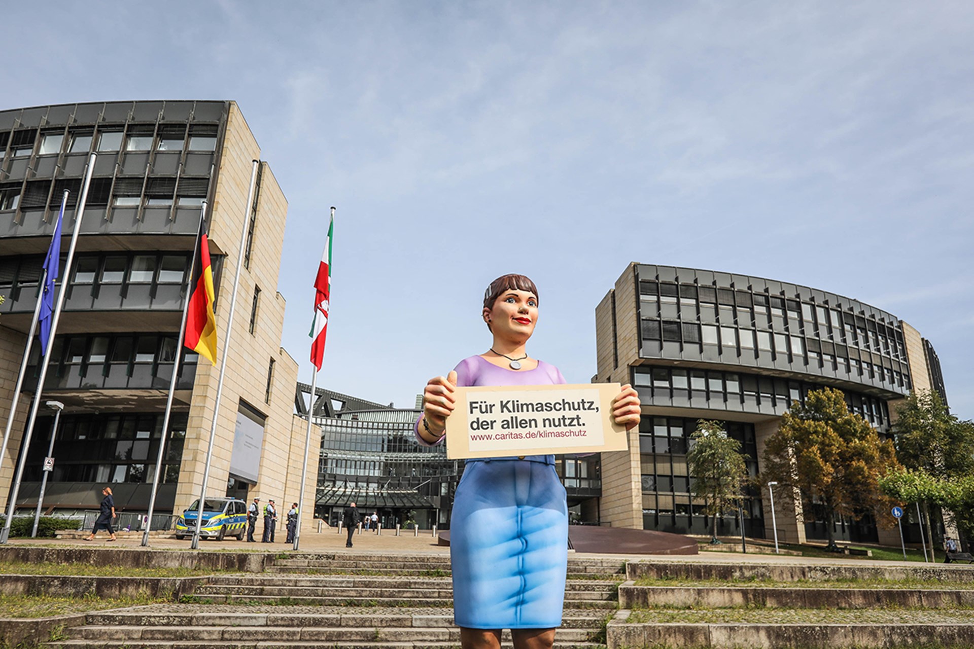 Die Fiberglasfigur 'Jenny', die vor dem Düsseldorfer Landtag steht und ein Schild mit einer URL und der Caritas-Kampagnentitel 'Für Klimaschutz, der allen nutzt.' trägt