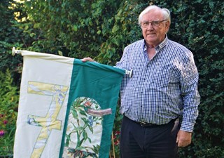 Lächelnder, älterer Mann, der in einem Garten steht und eine weiß-grüne Fahne mit einer eingestickten '70' hochhält