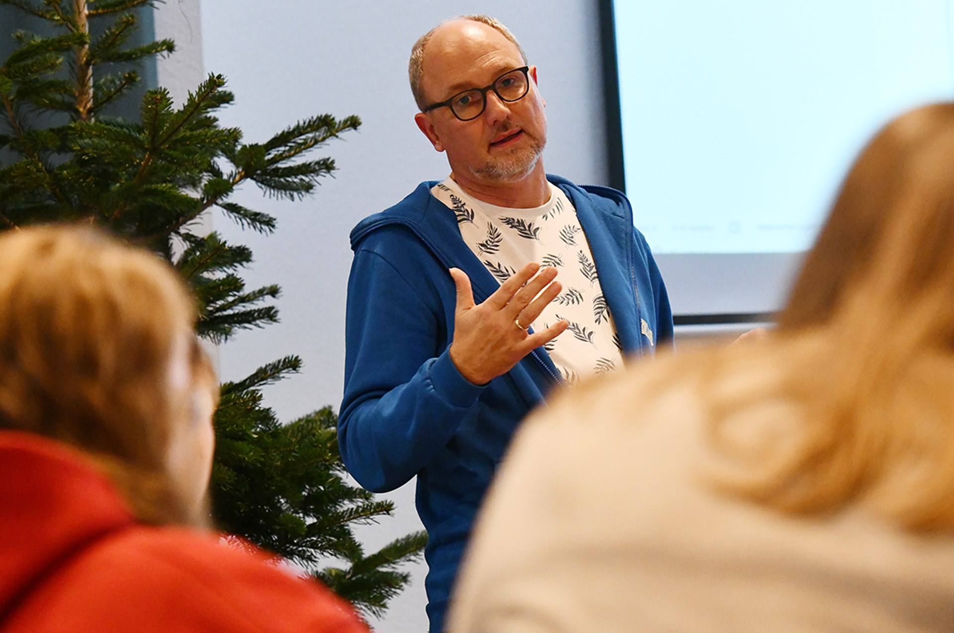 Ein Mann mit Brille, der ein bedrucktes T-Shirt und einen blauen Kapuzenpullover trägt, steht bei einem Schulung gestikulierend vor einer Videoleinwand und hält einen Vortrag. Links neben ihm ist ein Tannenbaum und im Vordergrund zwei weitere Personen zu sehen.