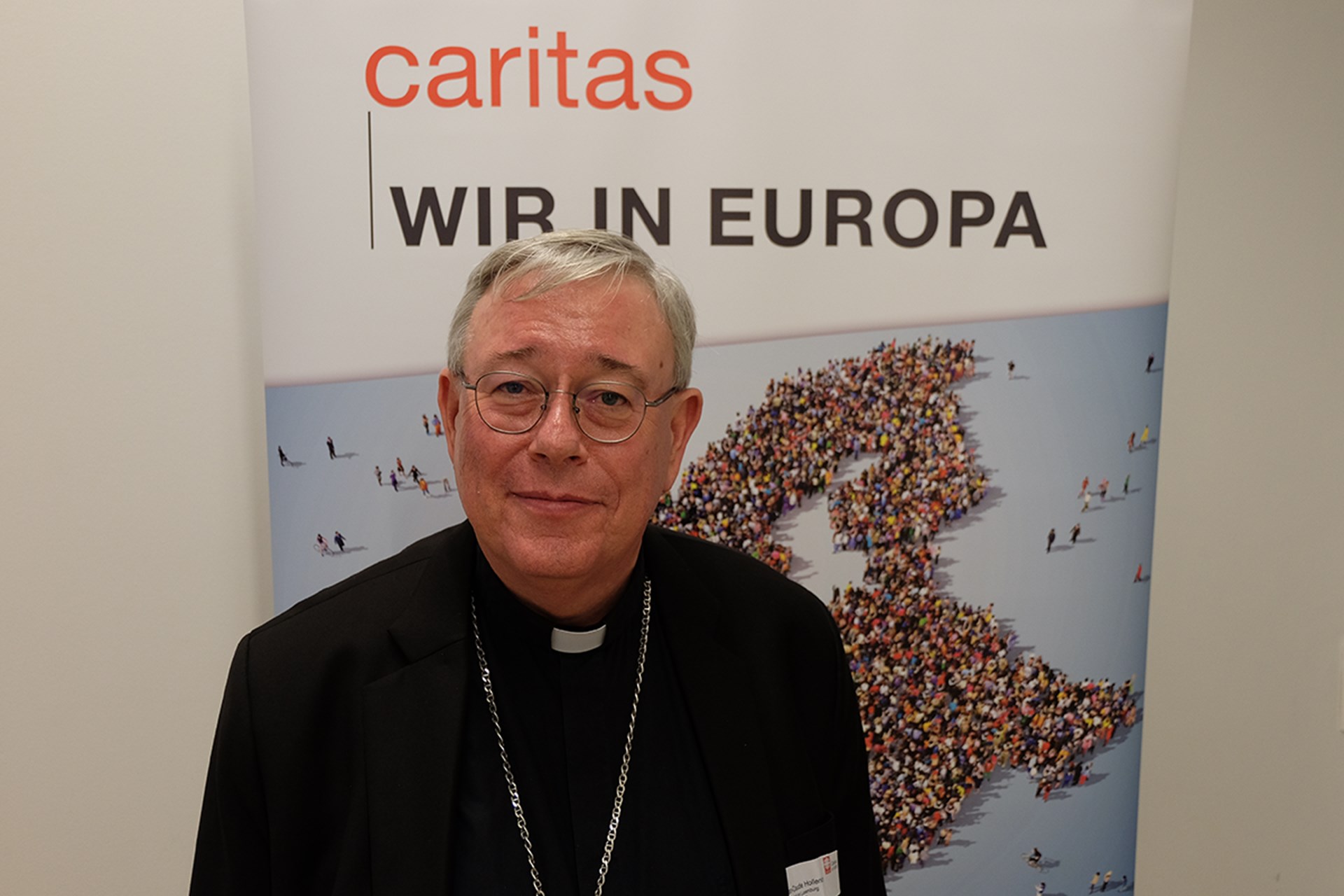 Ein Erzbischof, der auf dem Europaforum der Caritas vor einem Roll-up mit der Aufschrift 'caritas - Wir in Europa' steht