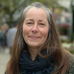 Lächelnde Frau mit langen, braun-grauen Haaren, die dunkle Kleidung trägt und sich in der Fußgängerzone der Essener Innenstadt befindet