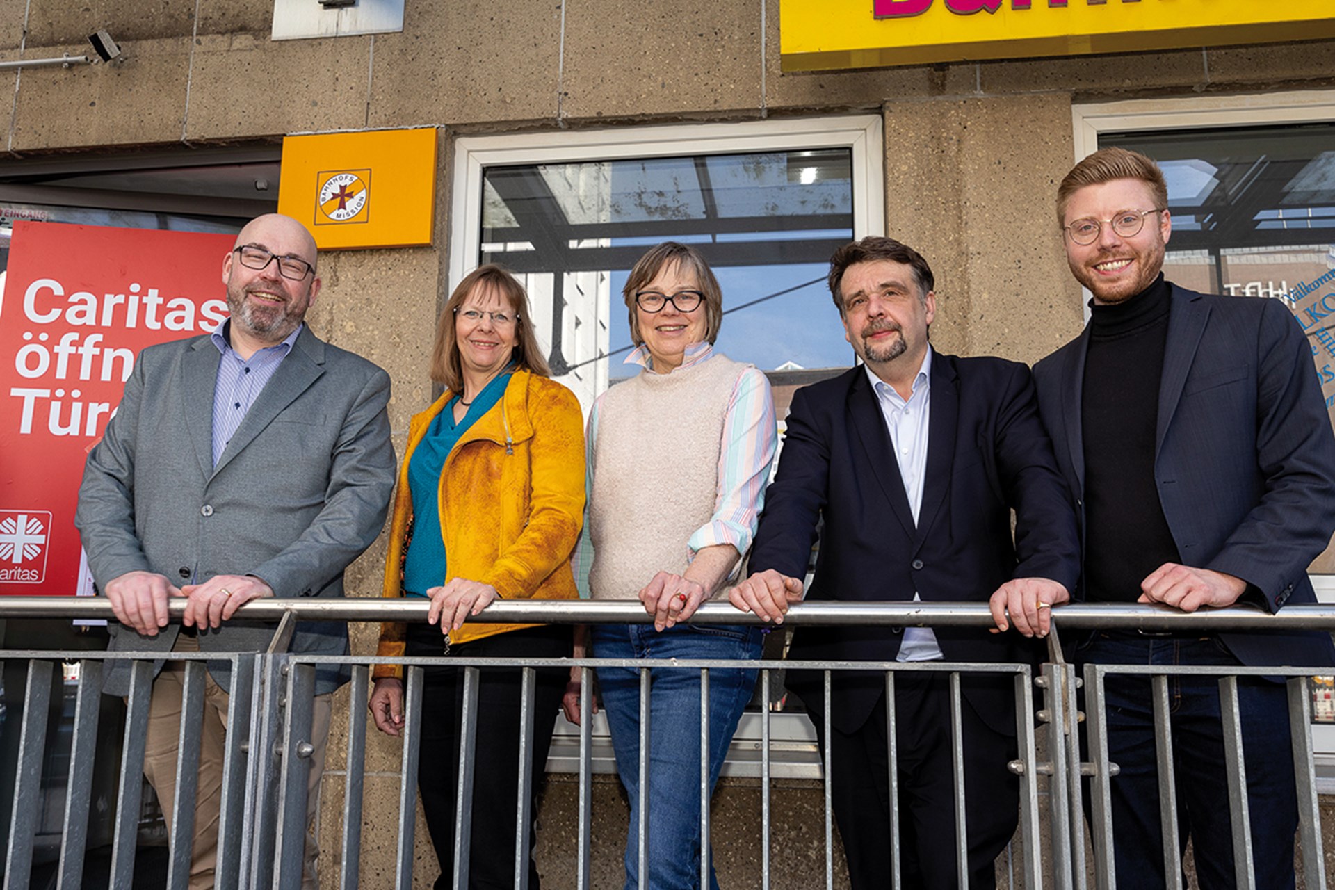 Eine Gruppe von drei Männern und zwei Frauen, die vor dem Eingang der Bahnhofsmission Essen an einem Geländer stehen