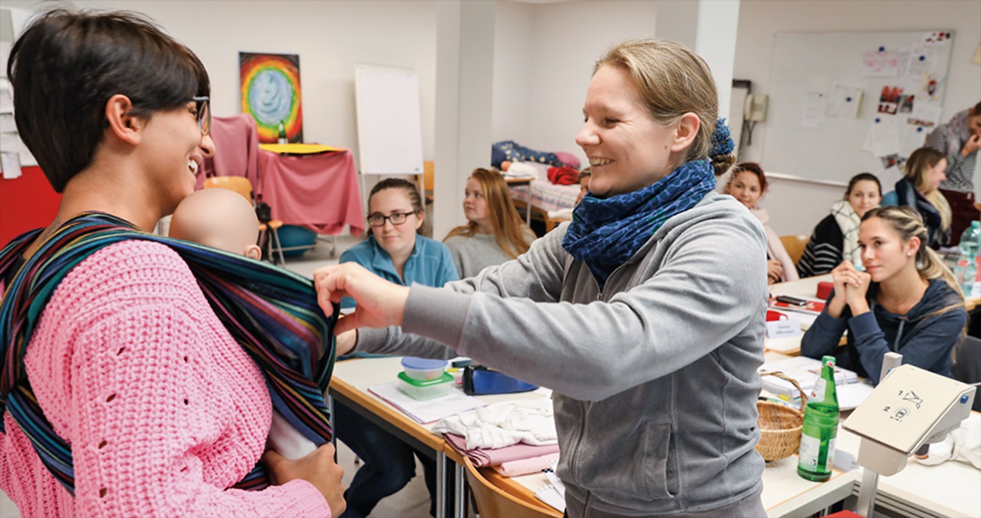 Einige Frauen sitzten in einem Schulungsraum an Tischen und blicken auf eine Ausbilderin, die einer Schülerin den Einsatz eines Tragetuchs zeigt