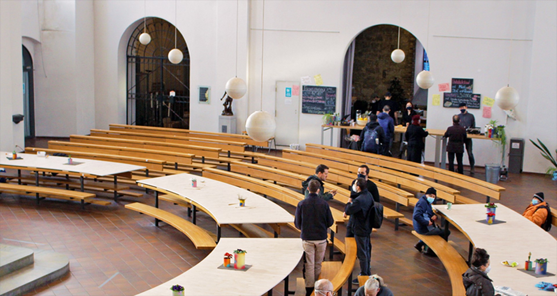 Der Innenraum der Kirche St. Peter in Aachen mit einigen Personen und einer aufgebauten Essensausgabe. Zwischen den Sitzbänken wurden mit Holzplatten provisorische Tische eingerichtet.