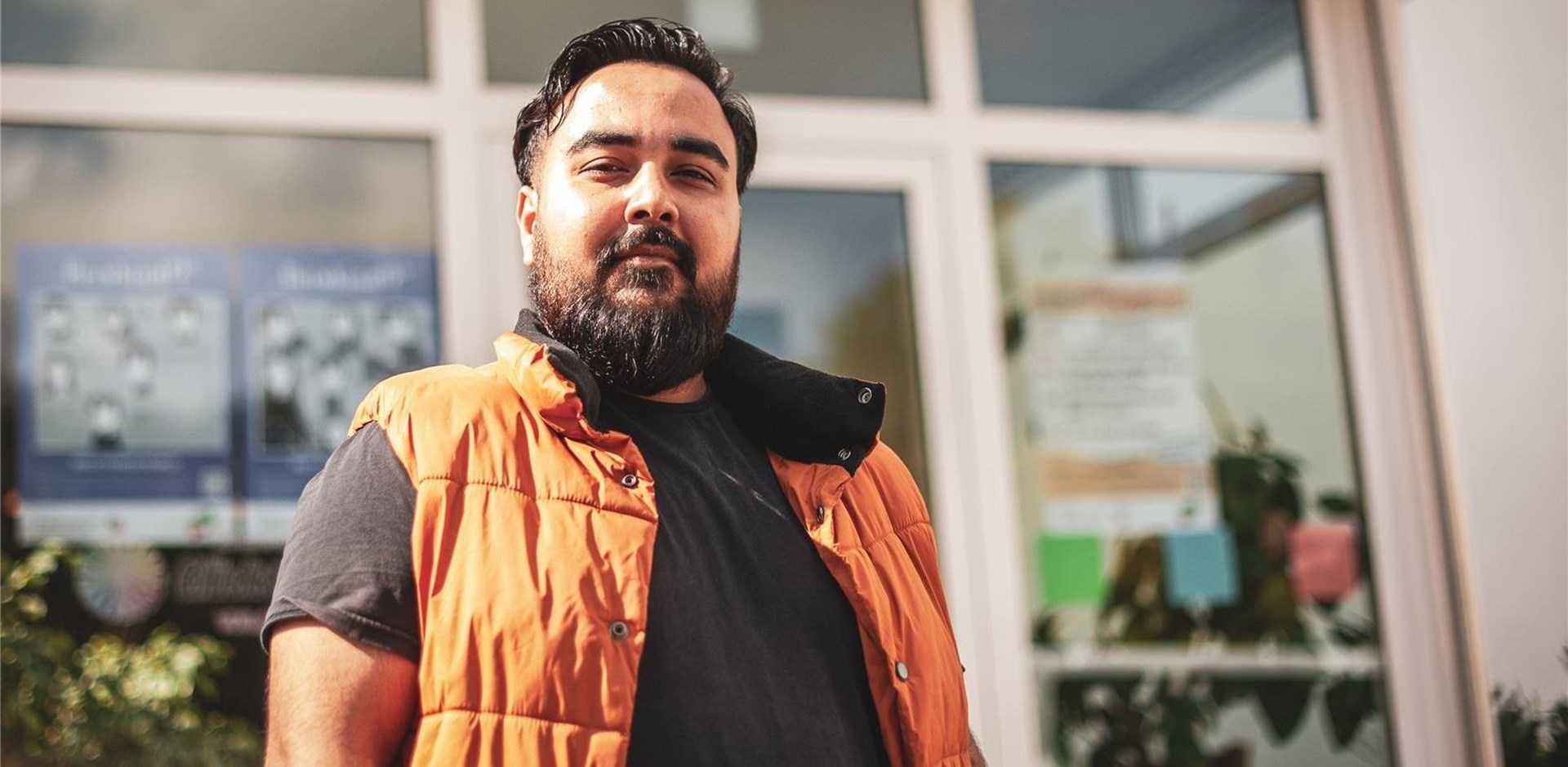 Ein junger Mann mit MIgrationshintergrund (dunkle Haare, dichter Bart) vor einem Gebäude