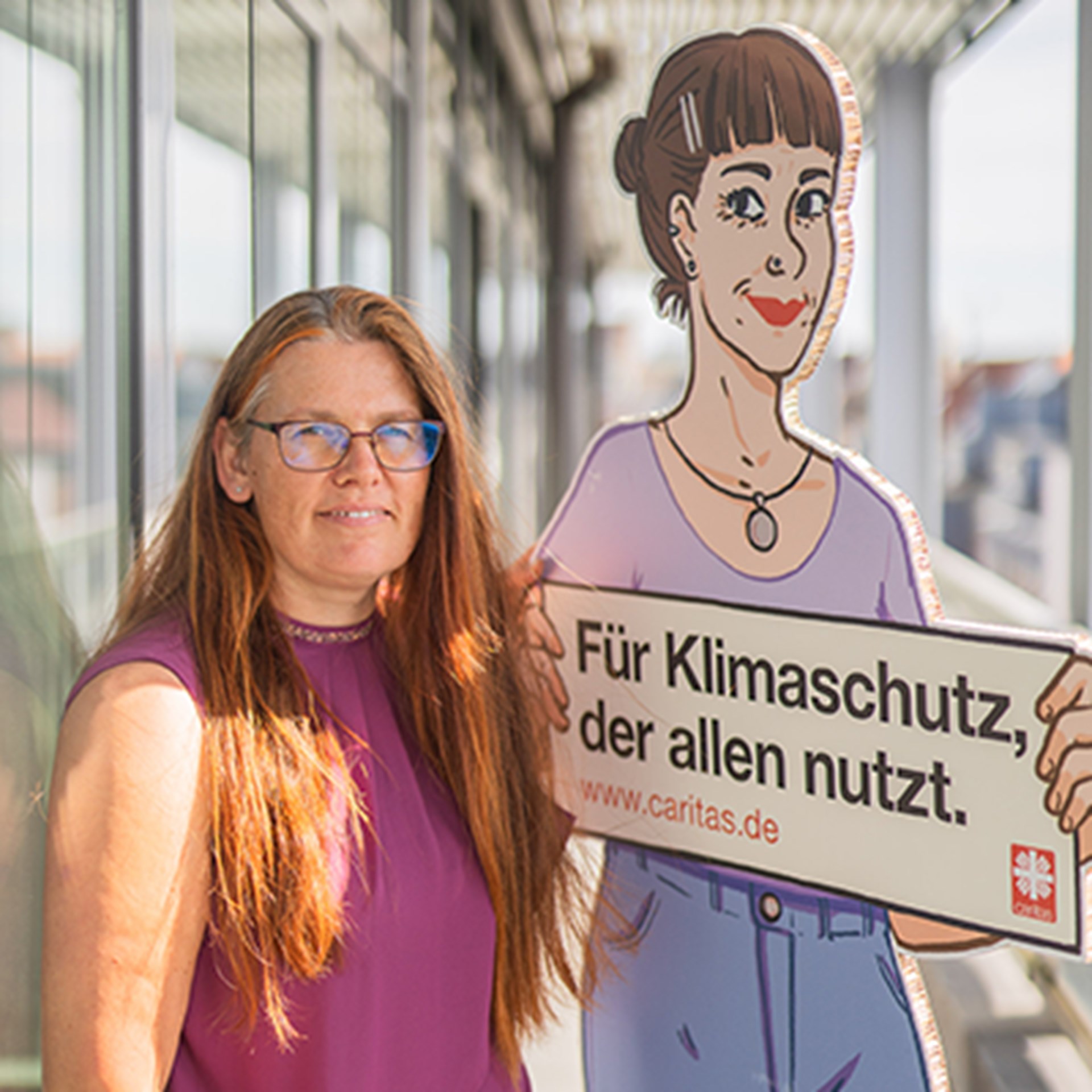 Lächelnde Frau mit langen, braunen Haaren und Brille, die ein violettes, ärmelloses Shirt trägt und in einem Laubengang steht. Neben ihr steht ein Pappaufsteller von 'Jenny' aus der Caritas-Jahreskampagne 2024 'Für Klimaschutz, der allen nutzt.'.