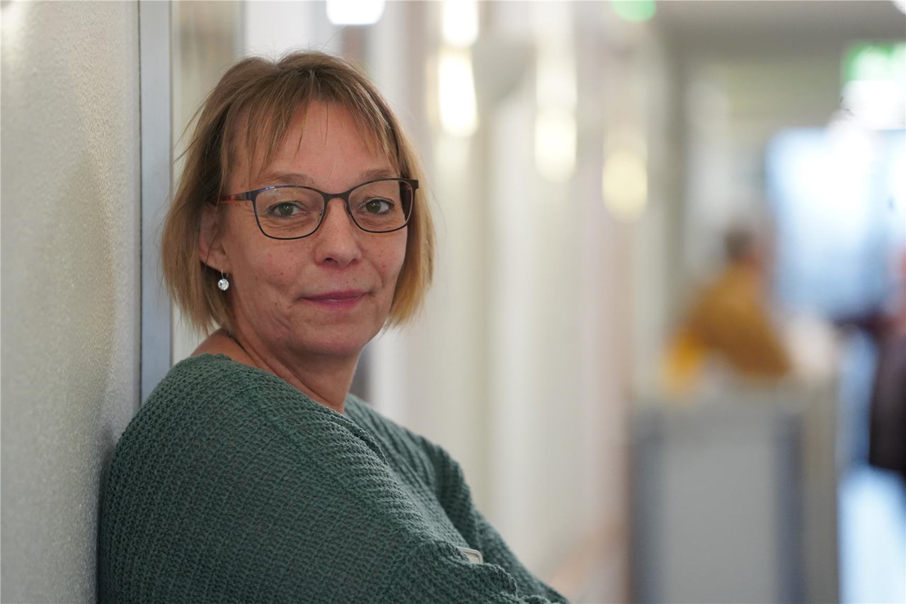 Lächelnde Frau mit blonden Haaren und Brille, die einen grünen Pullover trägt und im Flur einer Pflegeeinrichtung an einer Wand lehnt
