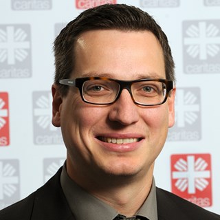 Lächelnder Mann mit braunen Haaren und Brille, der ein graues Hemd, eine graue Krawatte und eine schwarze Anzugjacke trägt. Er steht vor einem Roll-up mit grauen und roten Caritas-Logos.