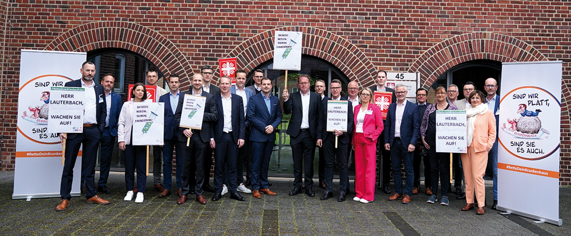 Ein Gruppenfoto mit mit Vertreter/innen der Caritas und der Kath. Kliniken im Bistum Münster, die eingerahmt von zwei Roll-ups mit Protestschildern vor der Geschäftsstelle des Diözesan-Caritasverbandes Münster stehen