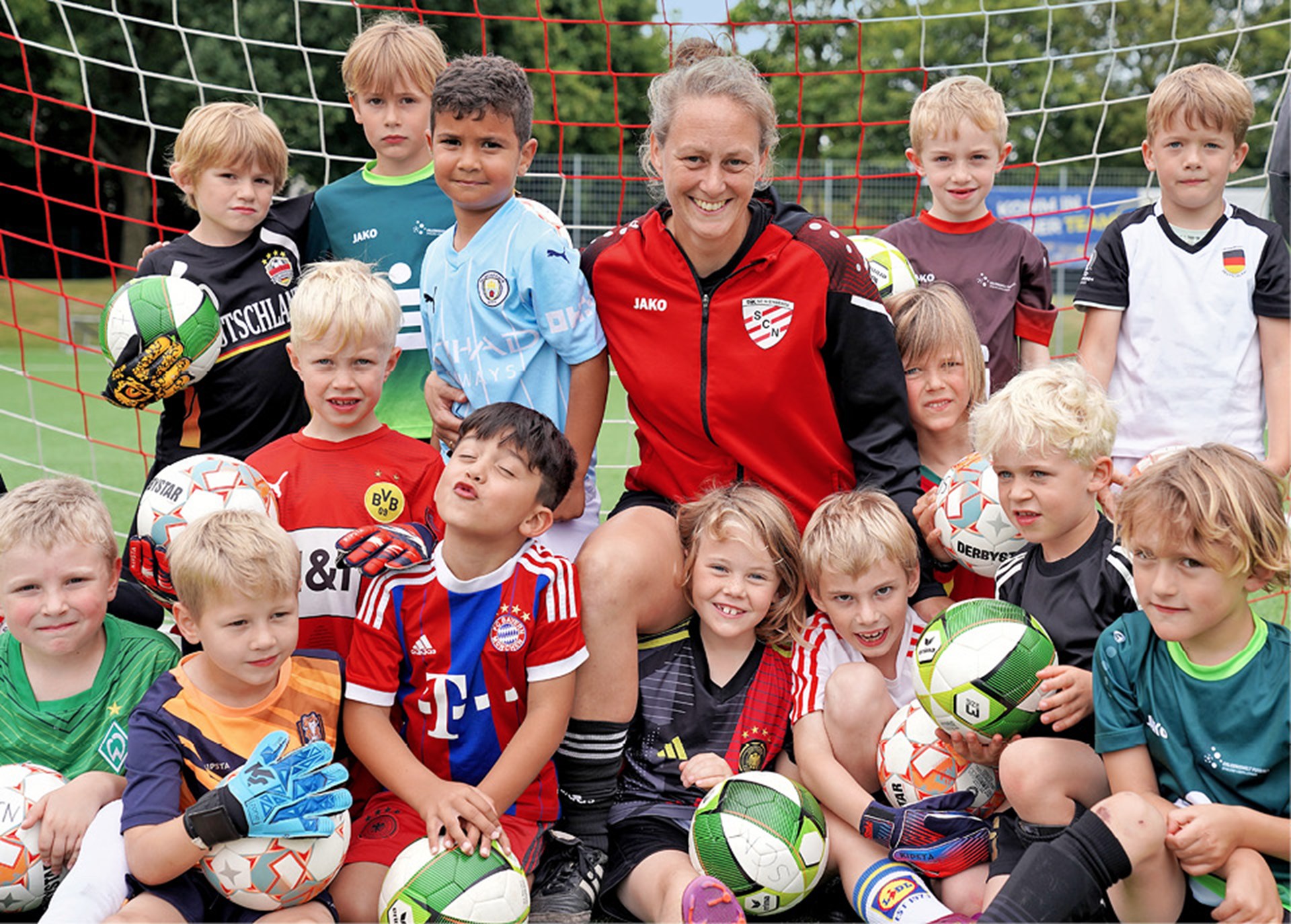 Gruppenfoto der U7 des DJK SC Nienberge mit ihrer Trainerin Liesa Marschall in einem Tor auf einem Fußballplatz des Vereins. Einige der Kinder haben einen Fußball in den Händen.