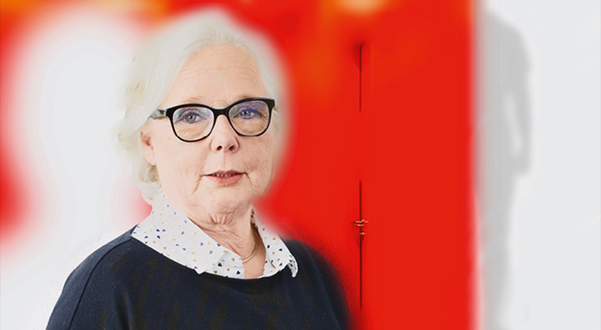 Ältere Frau mit weißen Haaren und schwarzer Brille, die ein weiß-gepunktetes Hemd und einen dunklen Pullover trägt. Sie wird vor einem rot-weißen Hintergrund dargestellt.