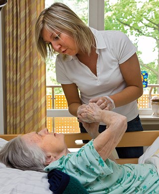 Eine Pflegerin steht an einem Krankenbett, in dem eine Seniorin liegt. Beide halten sich an den Händen und blicken sich in die Augen.
