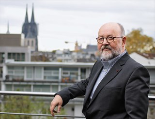Mann mit grauen Haaren, Halbglatze, Vollbart und Brille, der ein blau-gestreiftes Hemd mit einer grauen Anzugjacke trägt und auf einer Dachterrasse steht. Hinter ihm sind einige Häuser und der Kölner Dom zu sehen.