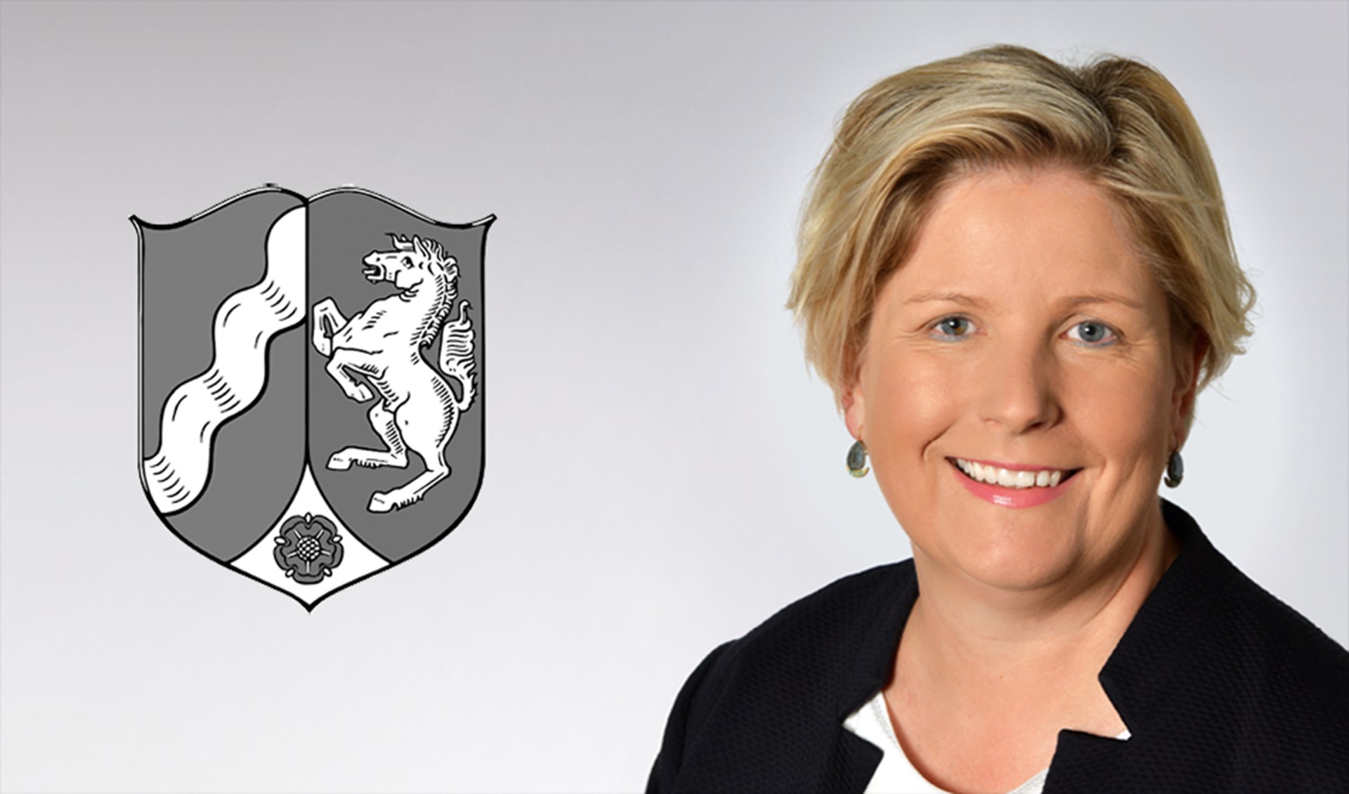 Lächelnde Frau mit blonden Haaren, die Ohrringe mit einer schwarzen Jacke trägt und sich auf der rechten Seite des Bildes befindet. Auf der linken Seite ist das Wappen des Landes Nordrhein-Westfalen in schwarz-weiß zu sehen.