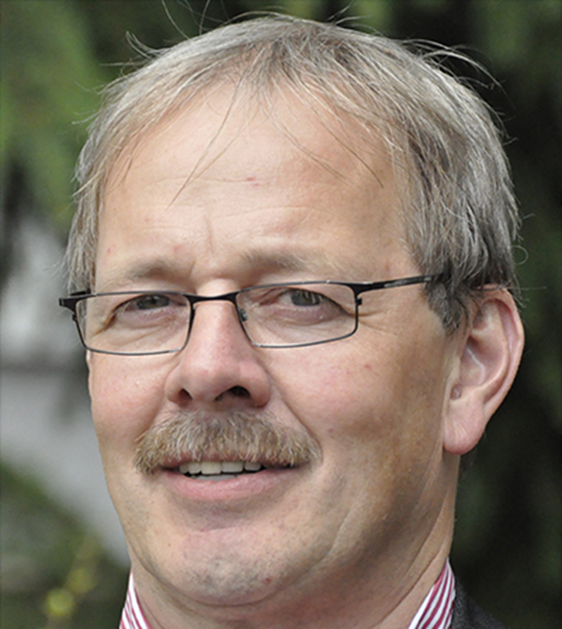 Lächelnder Mann mit grauen Haaren, Schnäuzer und Brille, der in einer Grünanlage steht