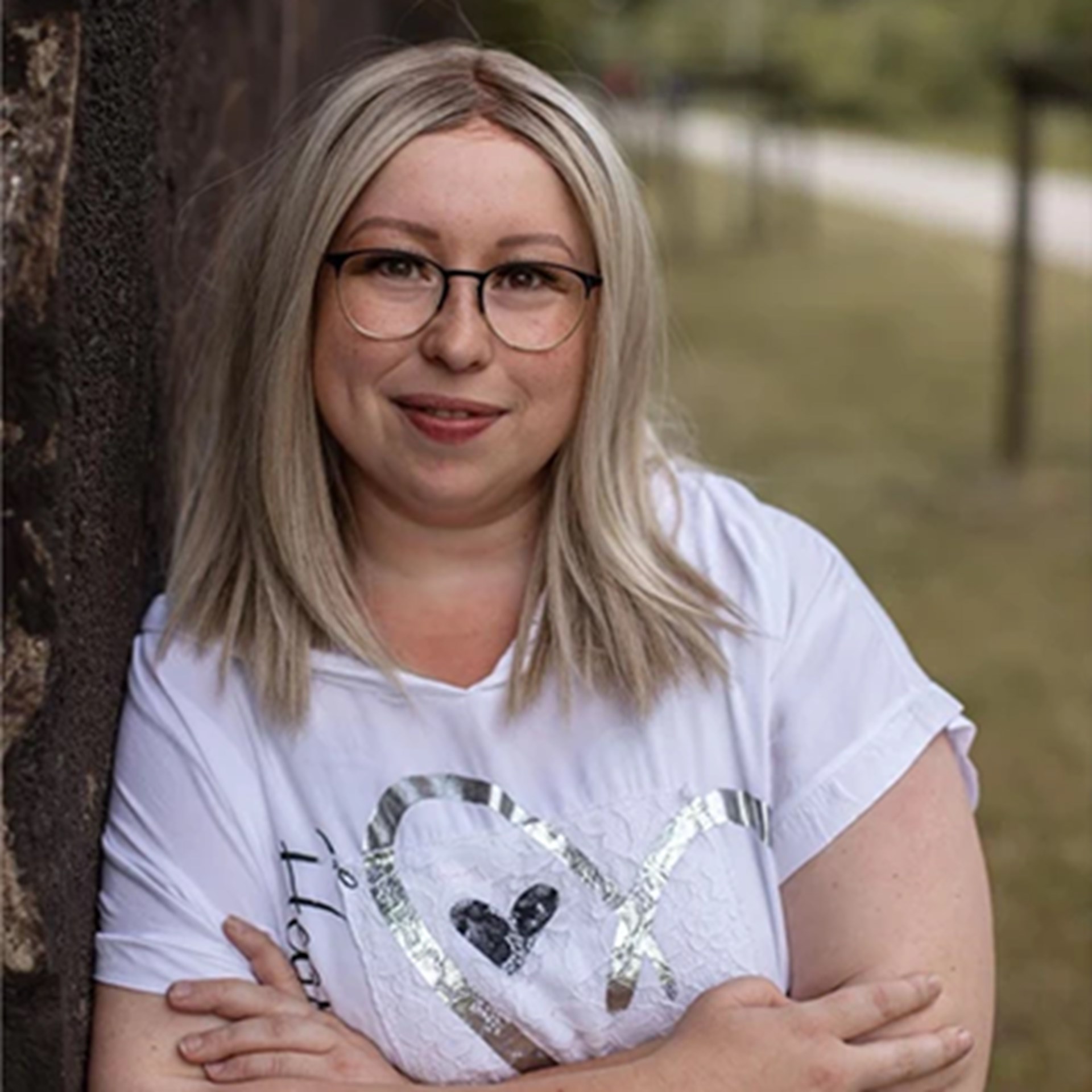 Lächelnde Frau mit blonden, schulterlangen Haaren und Brille, die ein weißes, gemustertes Shirt trägt und an einen Baum lehnt